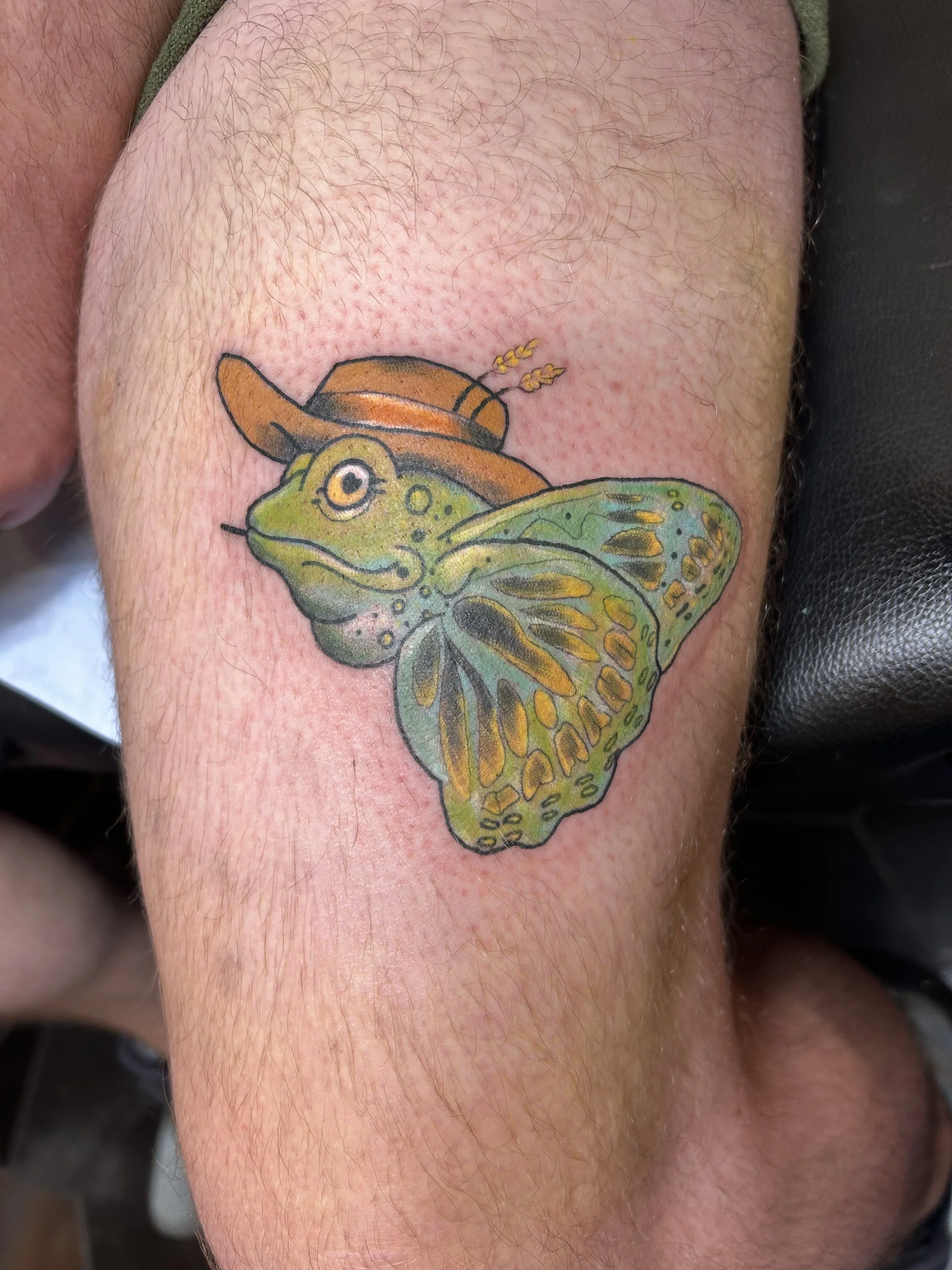 Tattoo of a green frog with wings wearing a brown cowboy hat and a small brown hat with wheat stalks on top, on a person's leg.