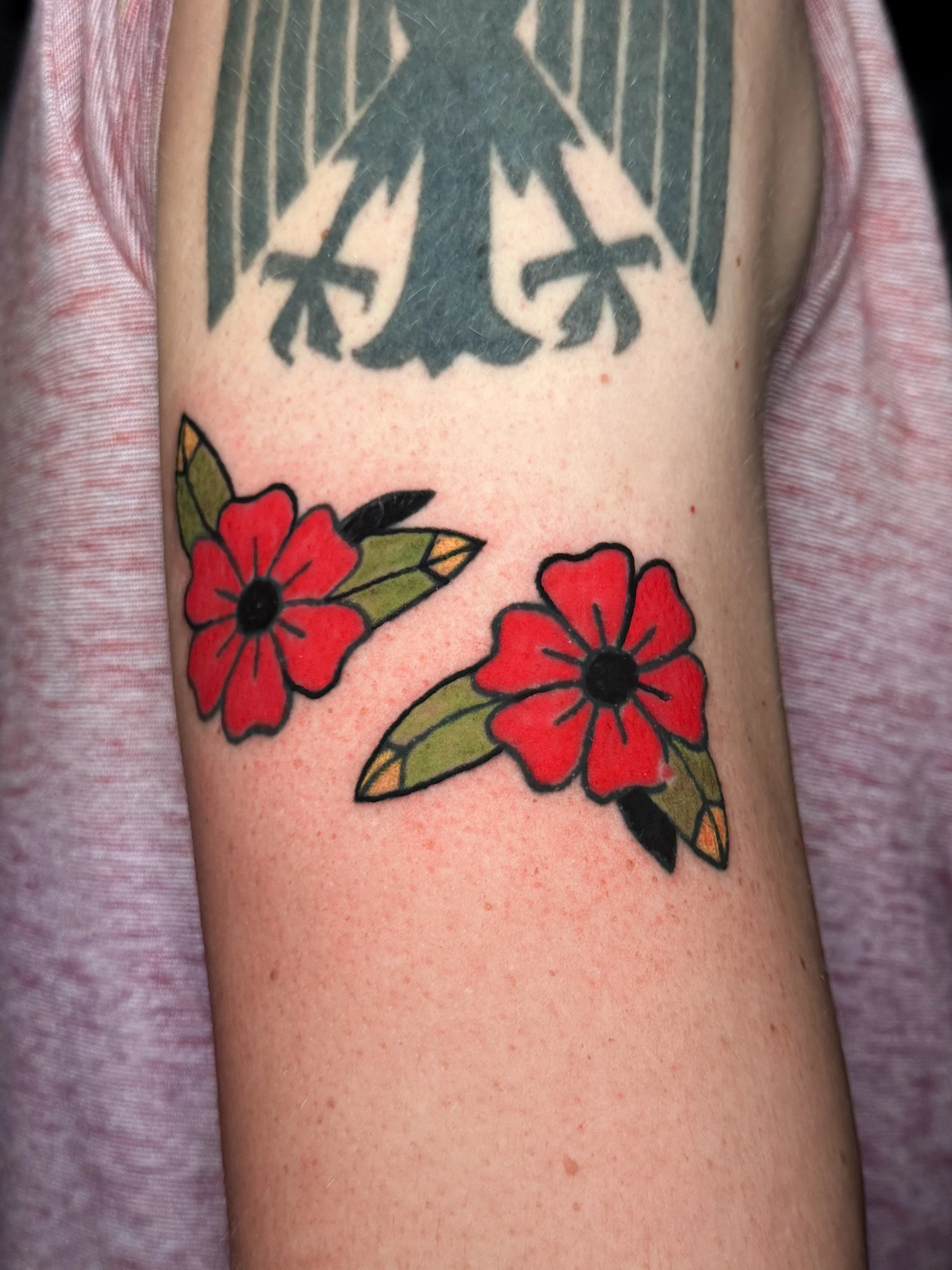 Close-up of a tattoo on a person's arm featuring two red flowers with green leaves, and a black and gray abstract design above the flowers.