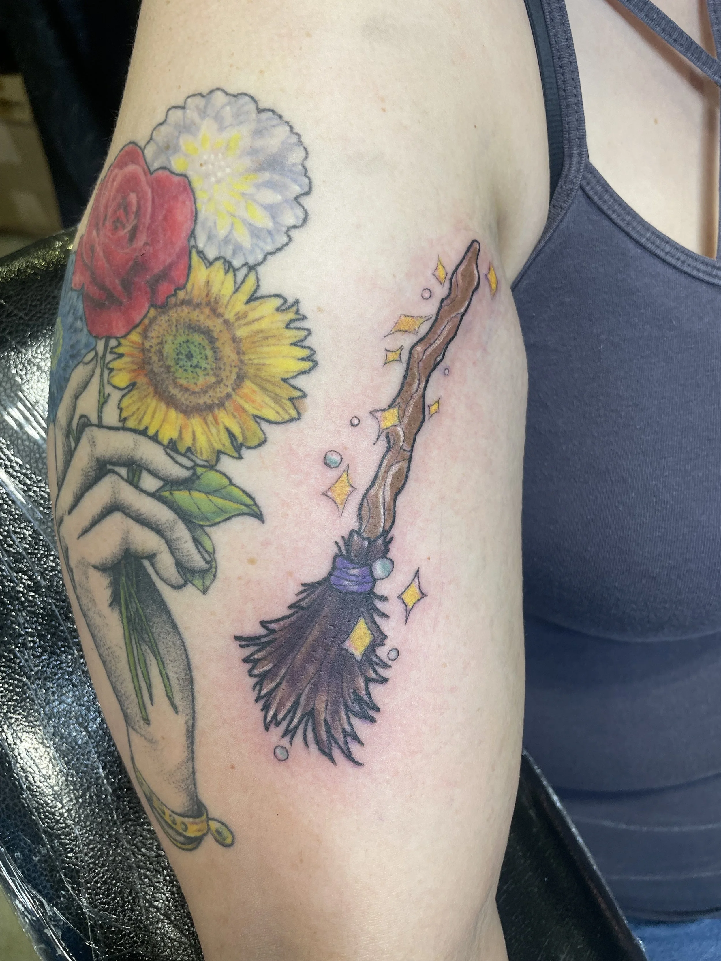 Tattoo of a hand holding a bouquet of colorful flowers, including a red rose, white flower, and yellow sunflower, with a witch broomstick surrounded by sparkling effects.