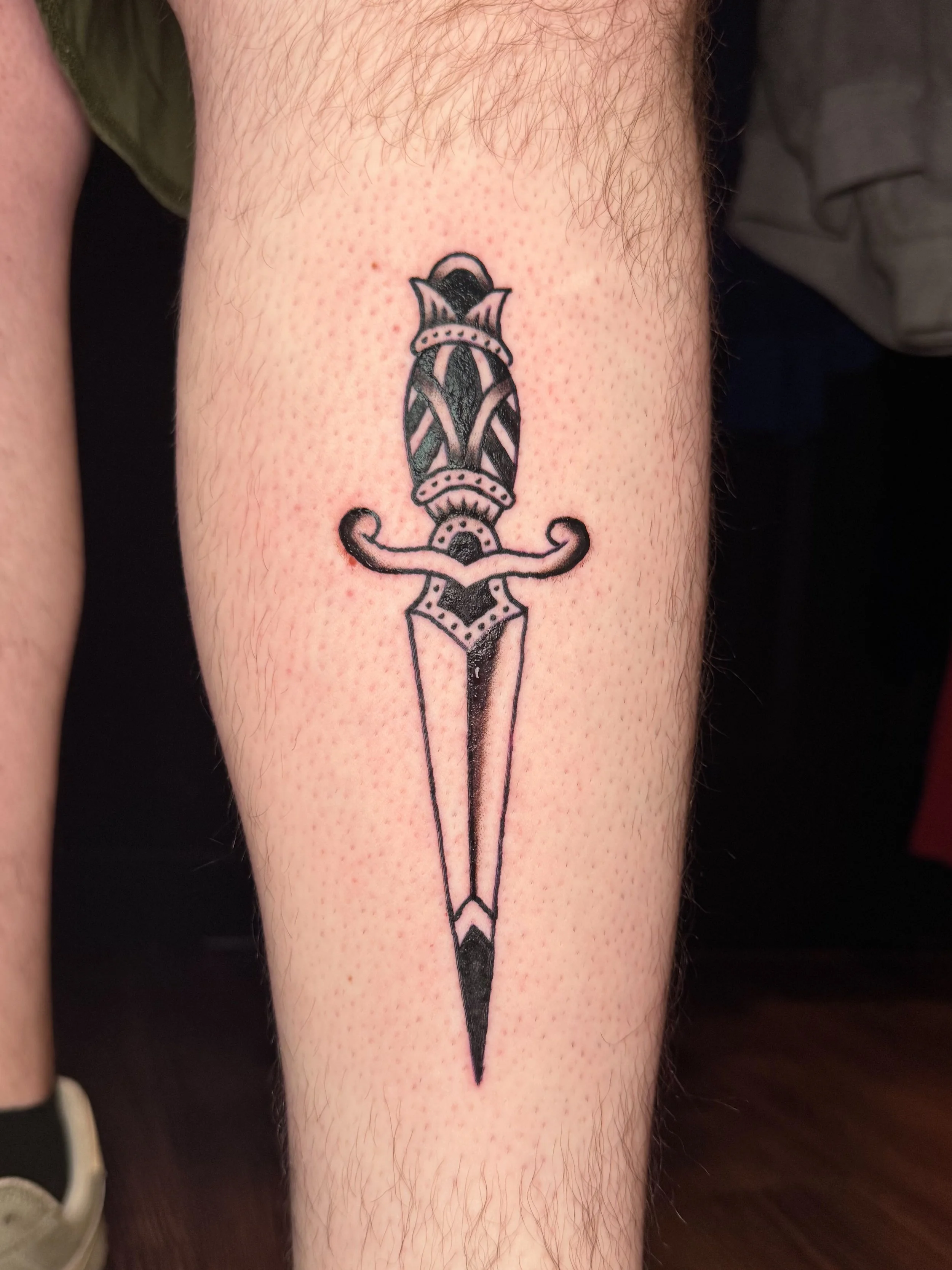 A tattoo of a medieval-style dagger with ornate details on the blade and hilt, inked on a person's lower leg.