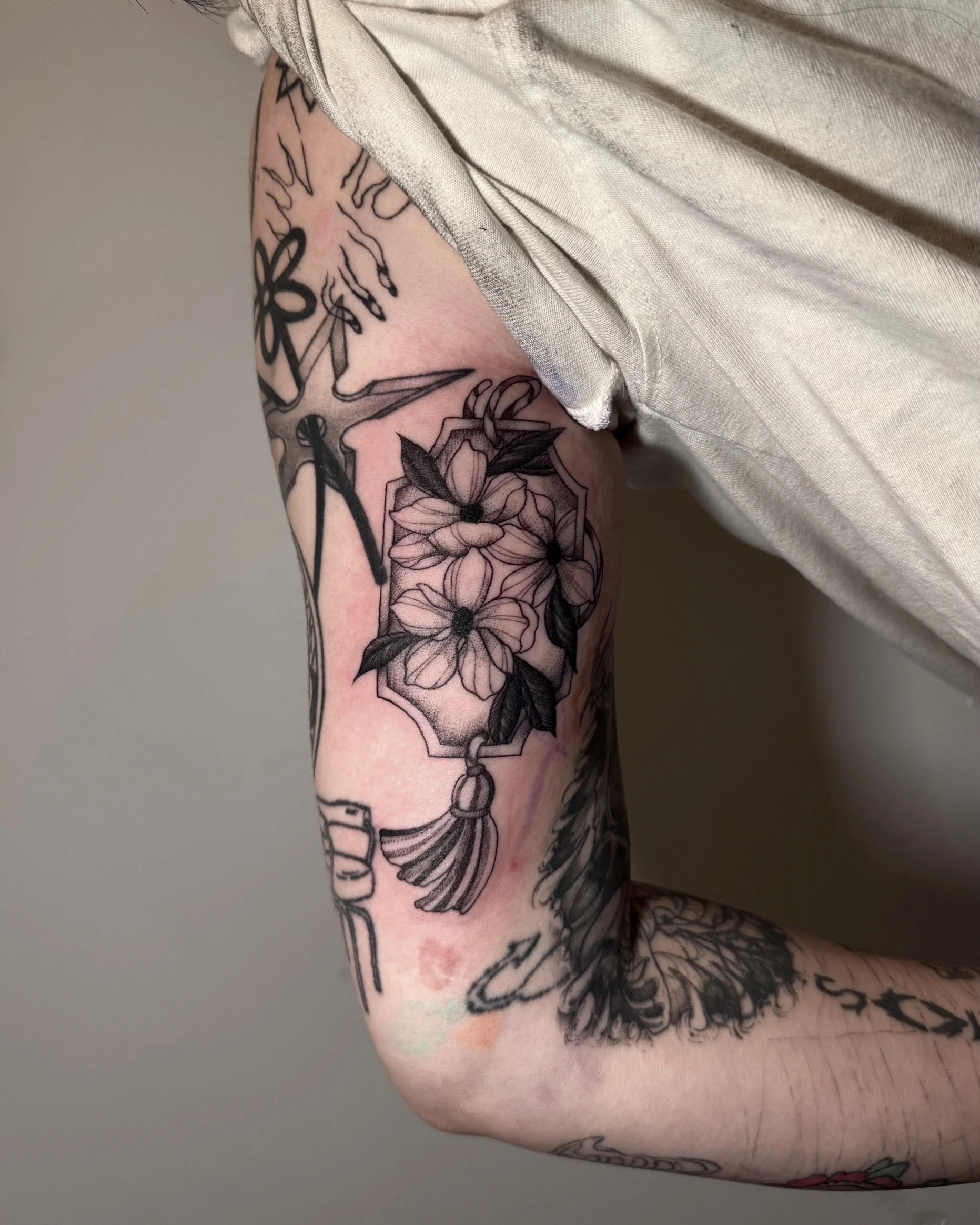 Black tattoo of a floral design with leaves and a hanging tassel on a person's upper arm.
