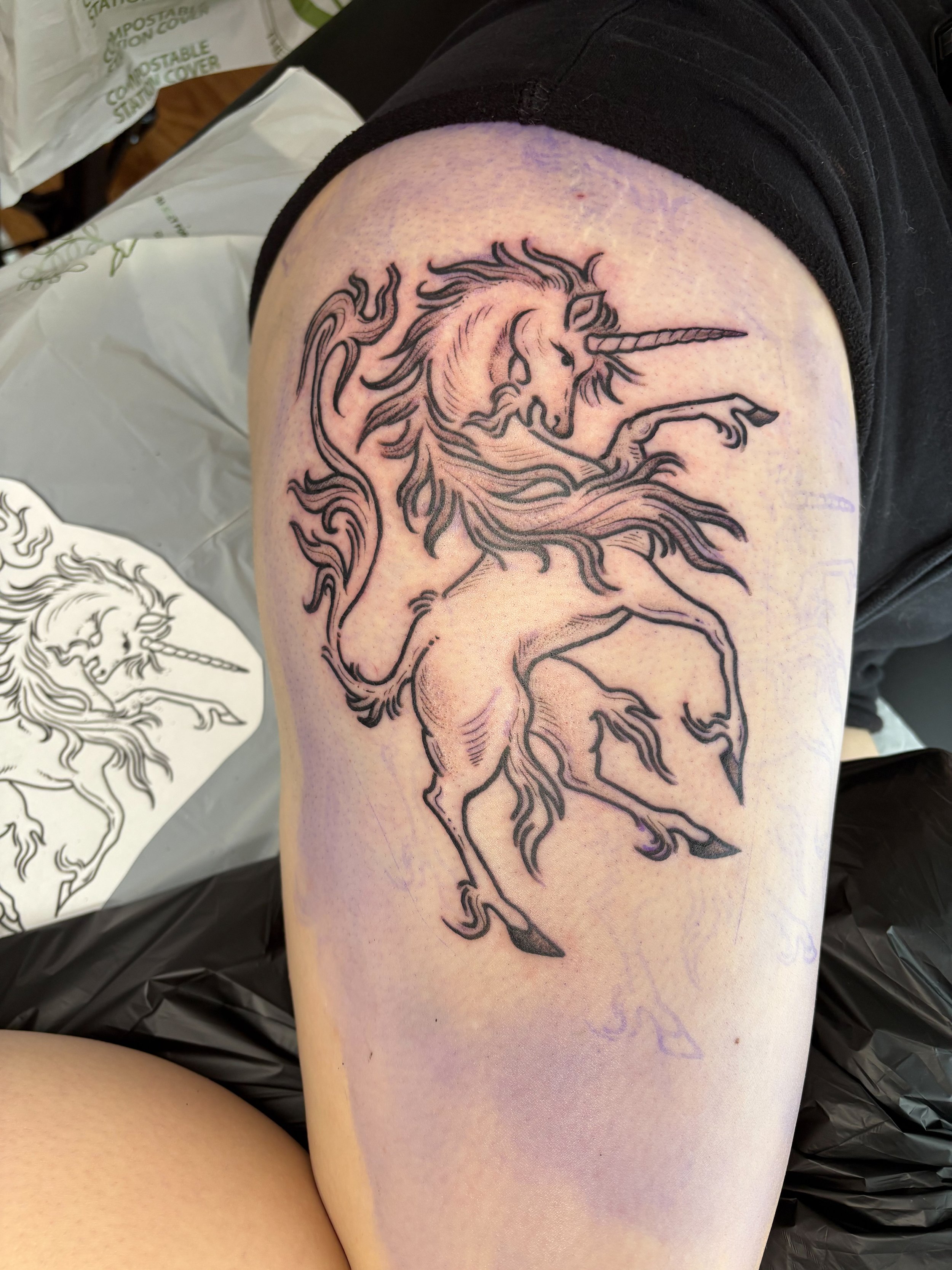 Unfinished black outline tattoo of a unicorn with a flowing mane and tail, with a twisted horn, on a person's upper arm.