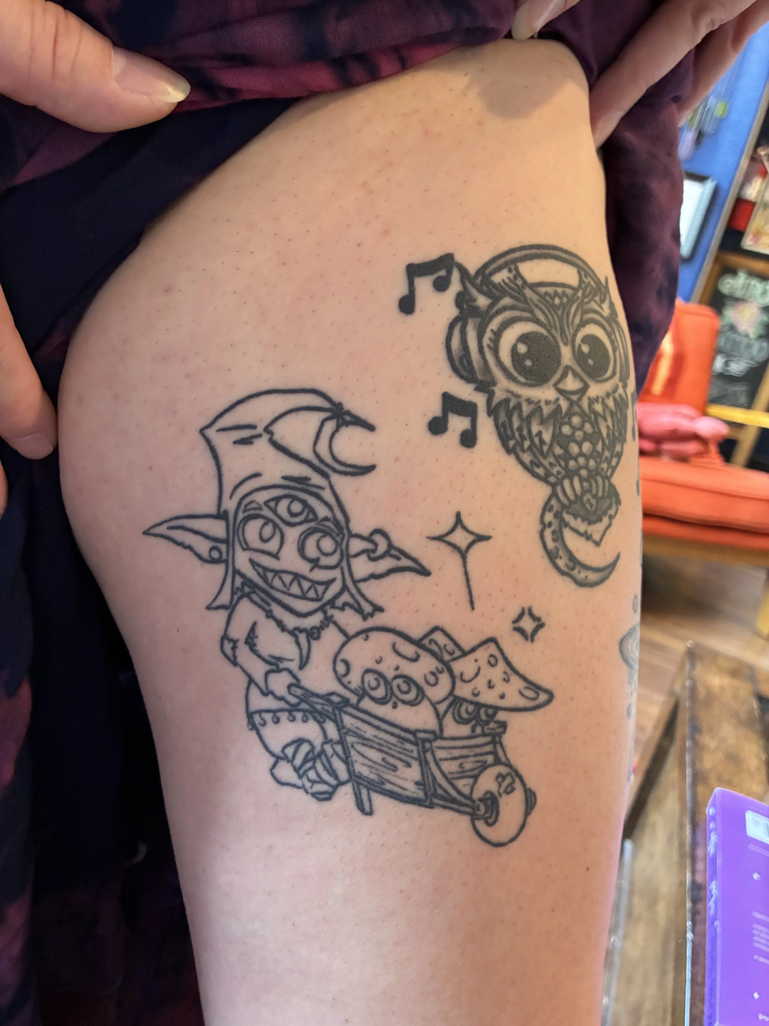 Tattoo of a cartoonish owl wearing headphones, with musical notes around it, on a person's thigh.