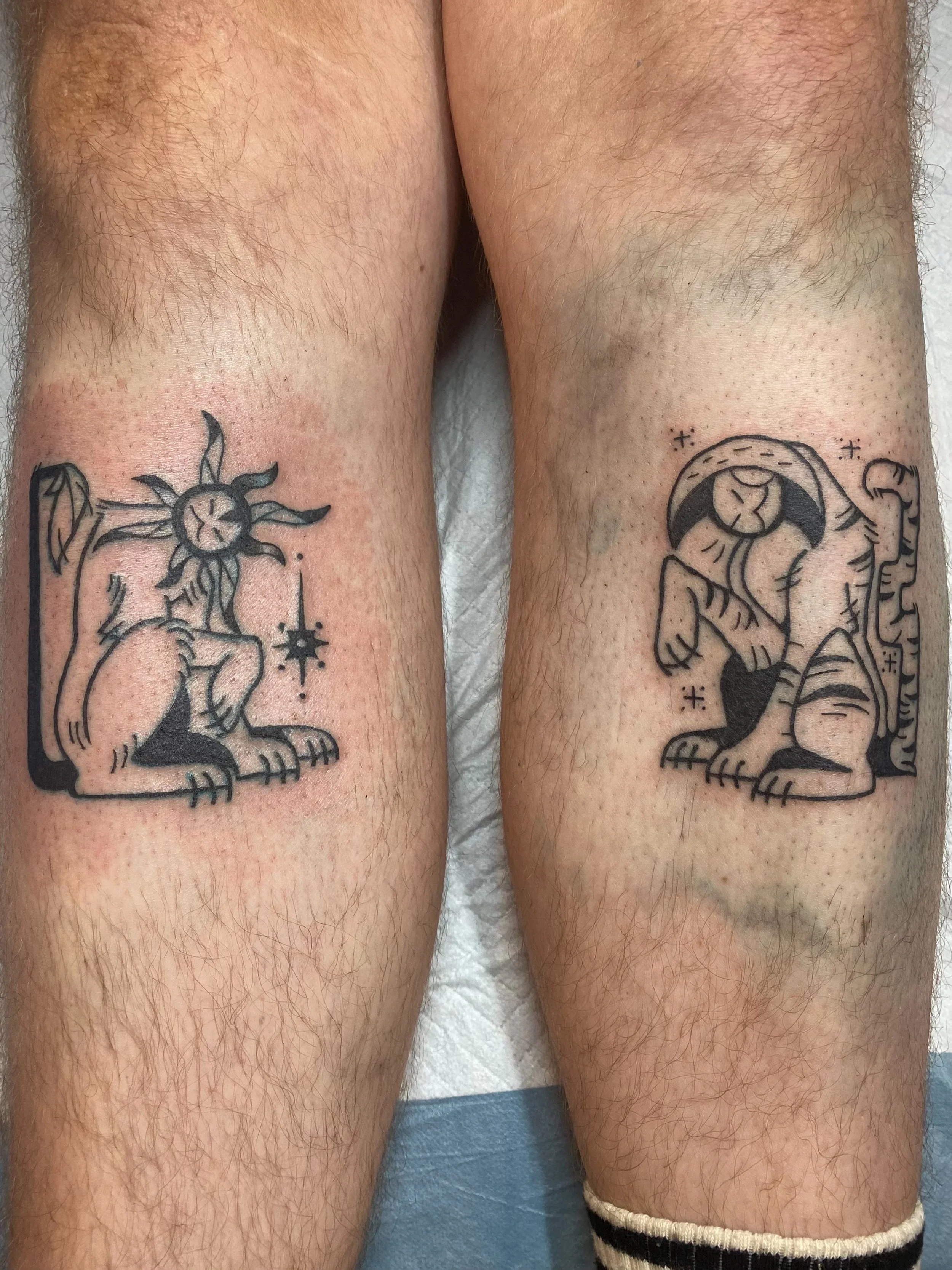 Close-up of two person's legs with tattoos, one featuring a lion with a crown and the other a sun with a face, on a blue surface