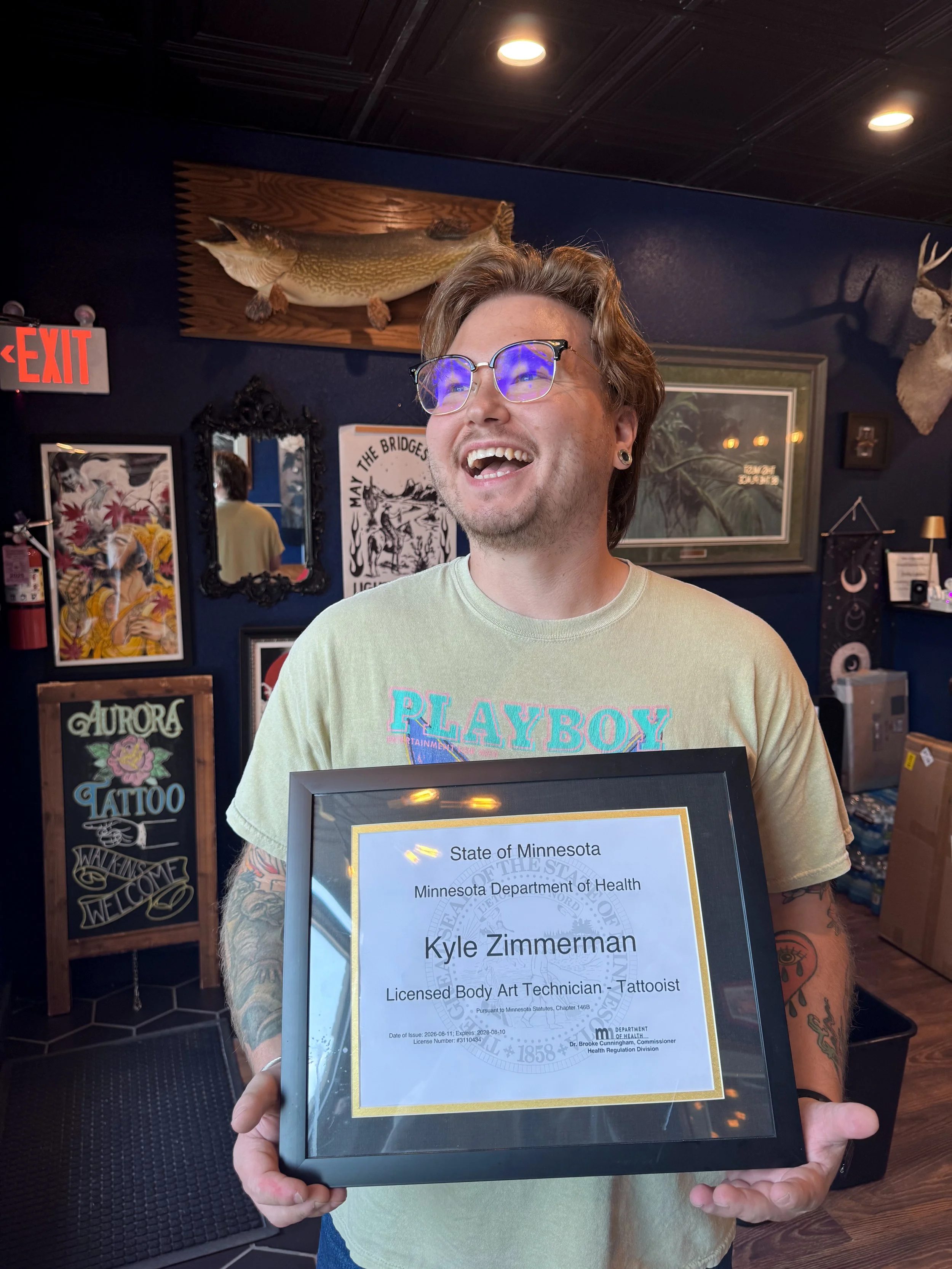 A smiling man with glasses holding a framed certificate from the Minnesota Department of Health. The man has tattoos and is in an indoor setting with dark blue walls decorated with various artworks and animal mounts.