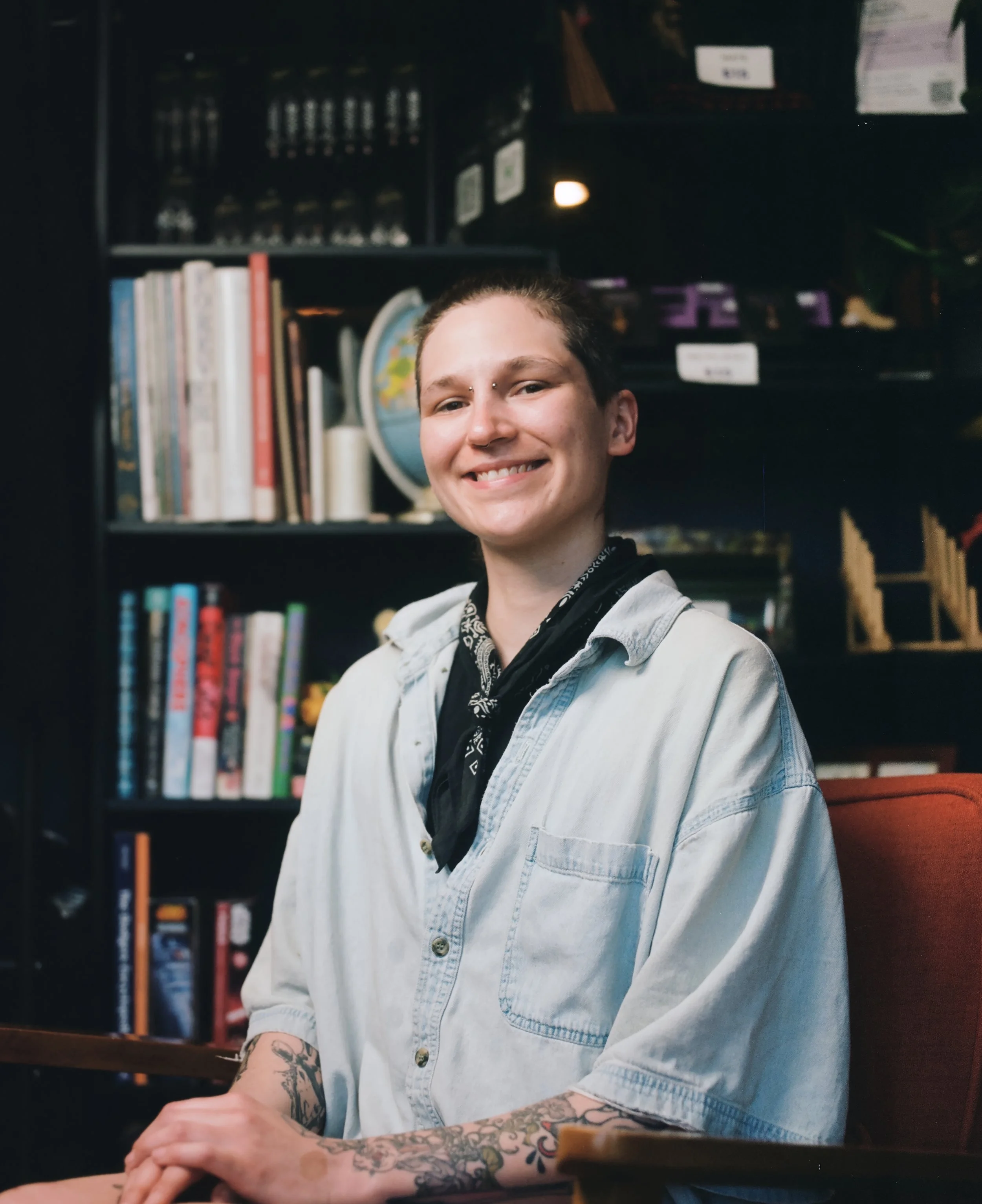 A smiling person with tattoos on their arms, wearing a light denim shirt and a black bandana, sitting at a table in front of a bookshelf filled with books, a globe, and various decorative items.