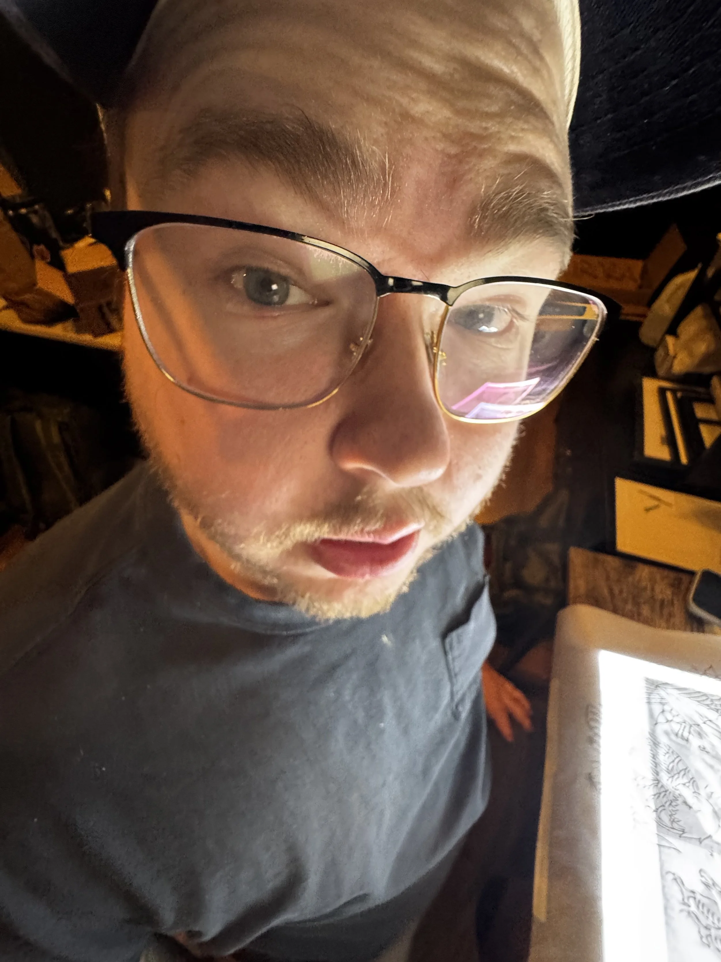 A young man with glasses looking up at the camera, with a tablet showing a black-and-white drawing in front of him.