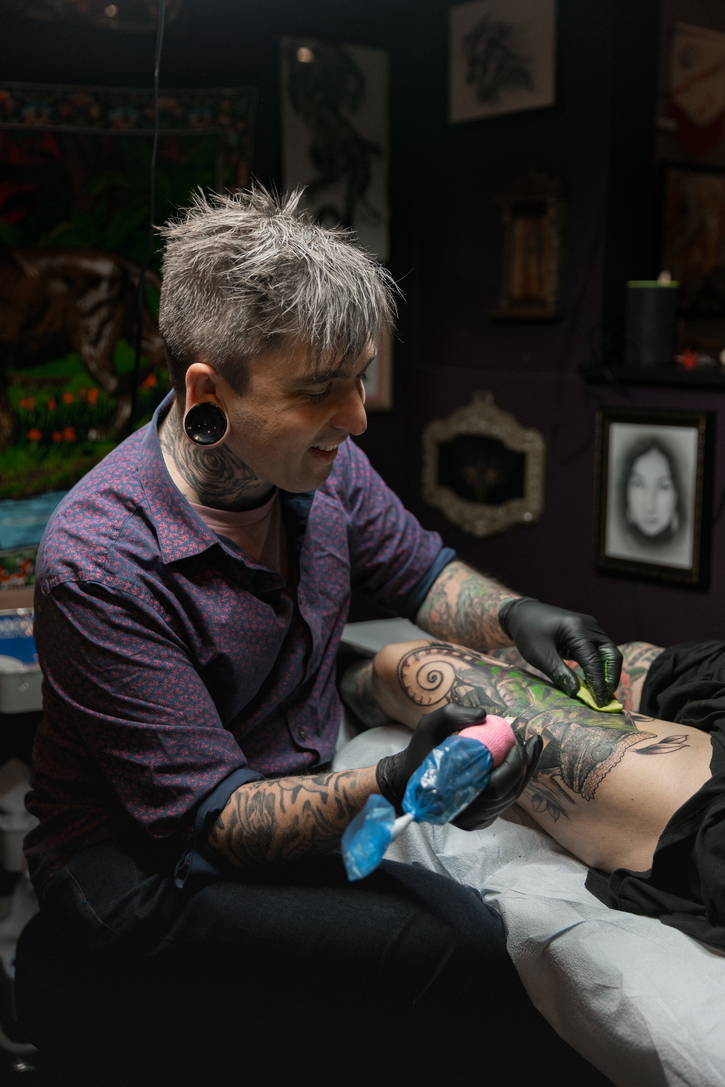 A tattoo artist with short gray hair, earrings, and tattoos on her arms, wearing black gloves, is tattooing a person's leg with a large tattoo.