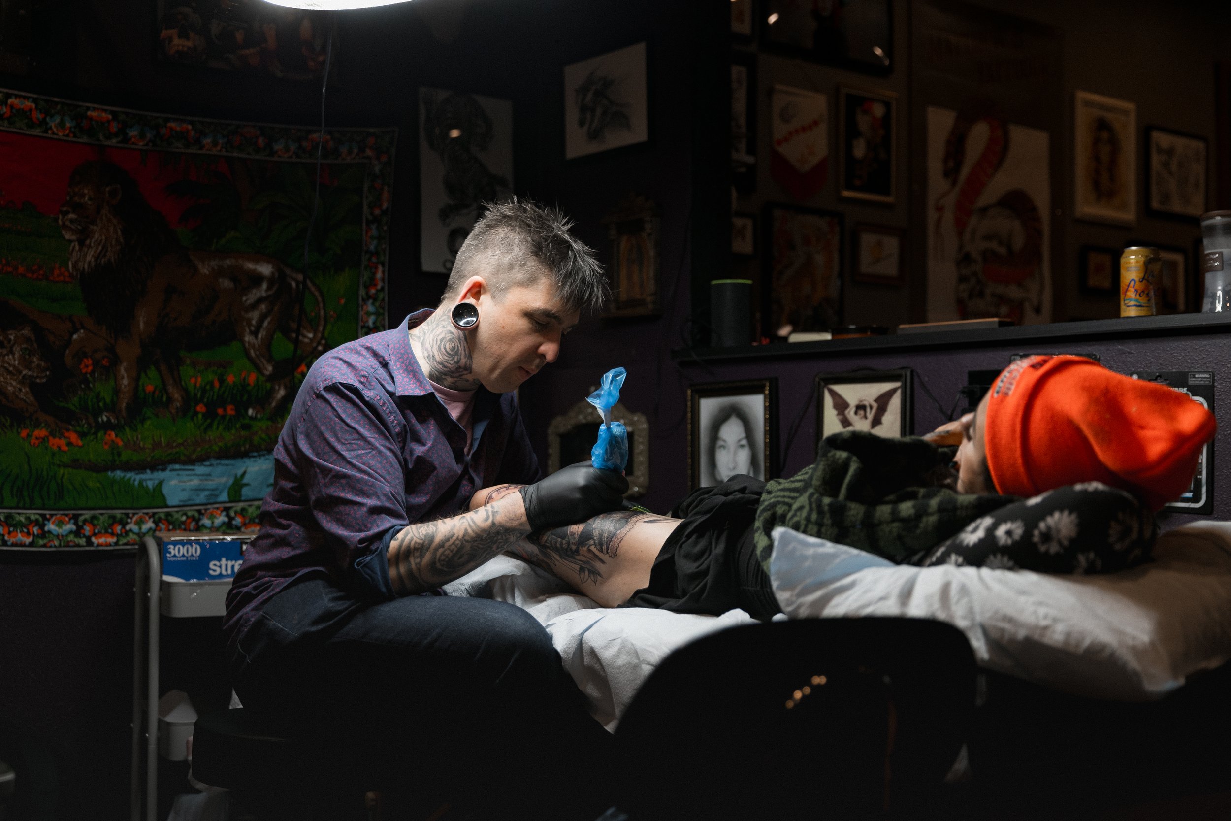 Tattoo artist giving a tattoo to a client lying on a bed, surrounded by dark walls decorated with framed art and a large tapestry of a lion, in a tattoo studio.