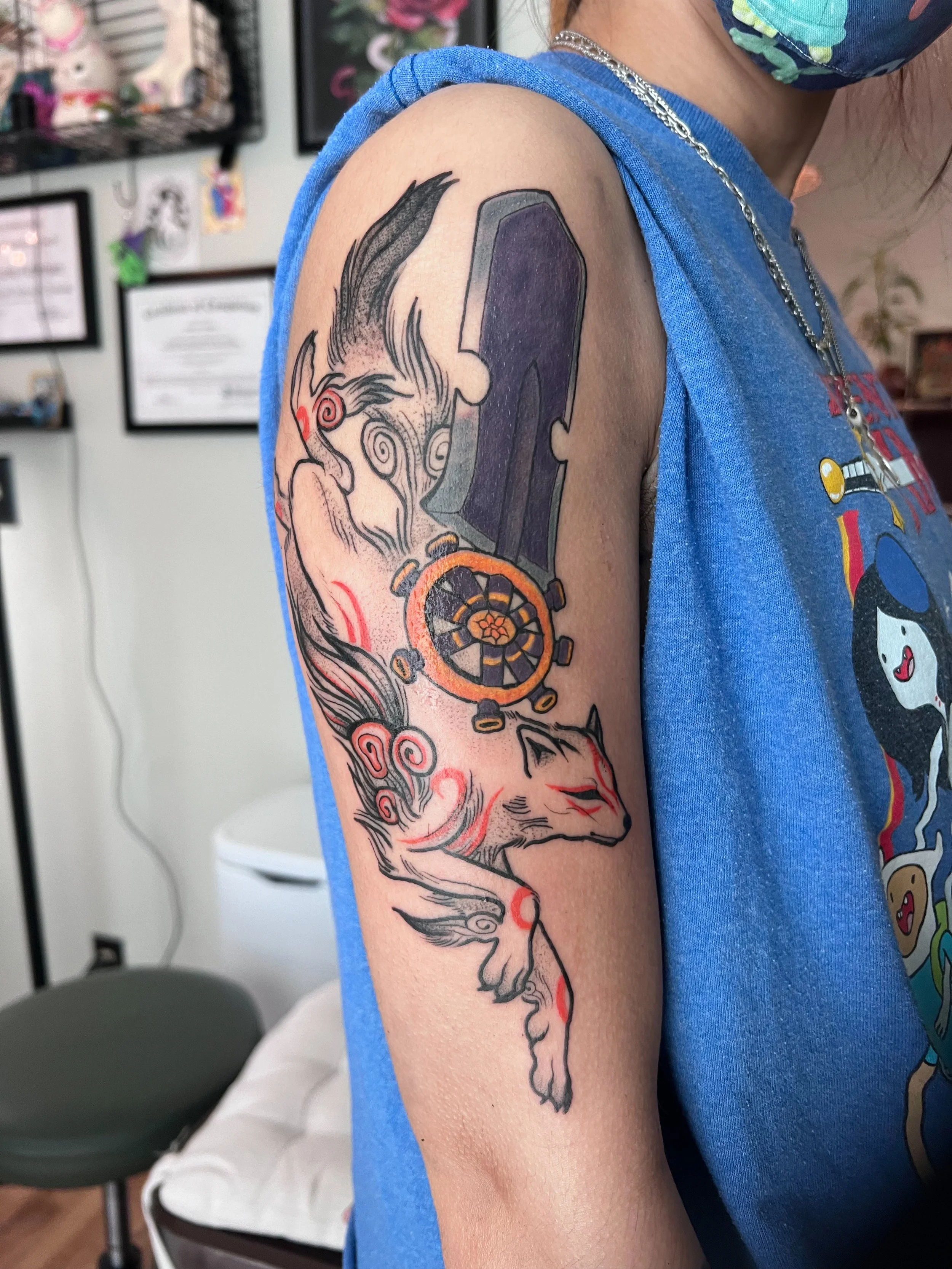 Tattoo of a fox with red markings and swirling patterns, a large sword, and a wheel or compass on the person's upper arm.