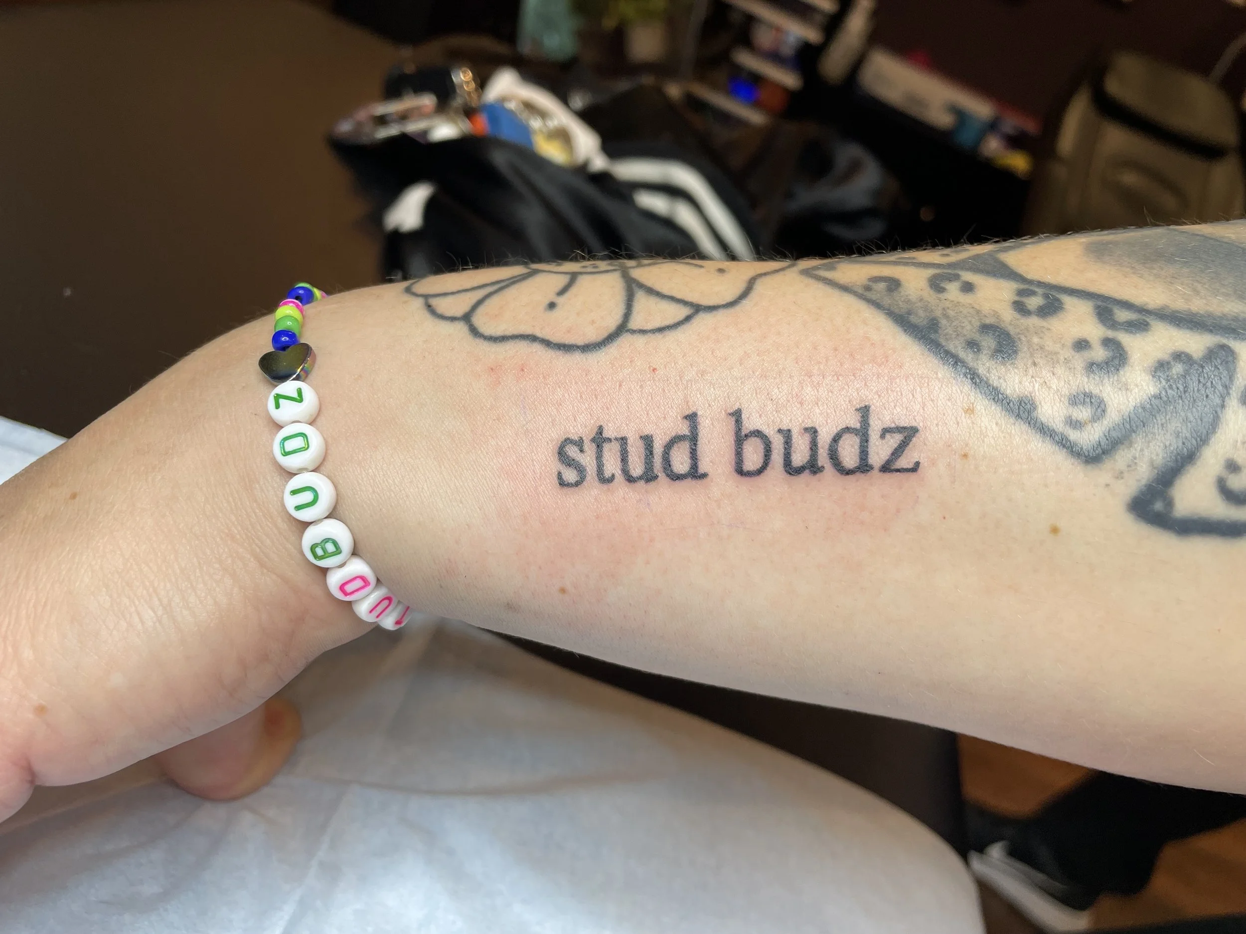 A person's arm displaying a tattoo that reads 'stud budz', alongside a colorful beaded bracelet with round letter beads spelling 'DUBUZO', and some additional tattoos in the background.