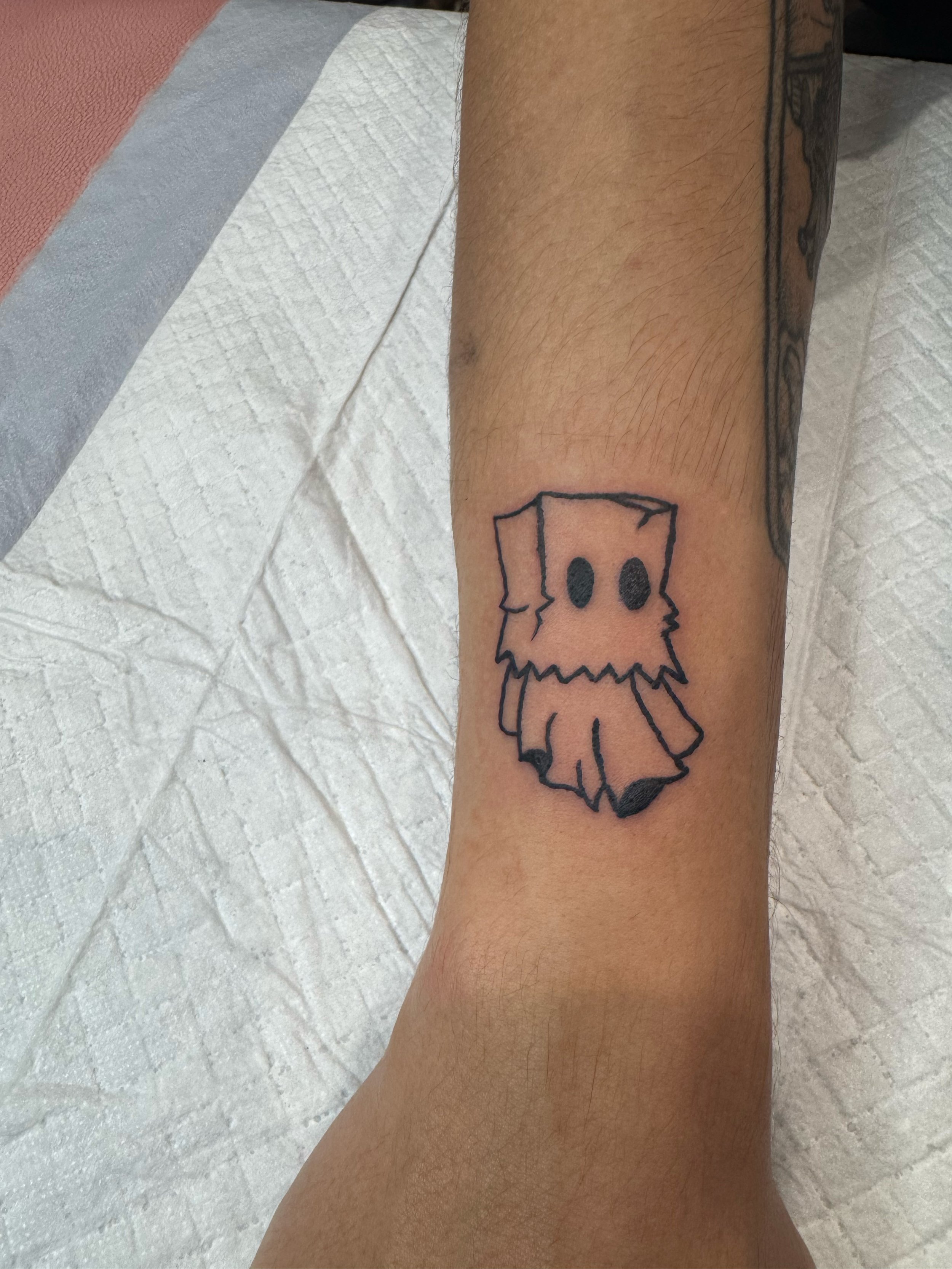 Tattoo of a cartoon-style ghost with black oval eyes and a tattered sheet covering its body, outlined in black ink on a person's forearm.