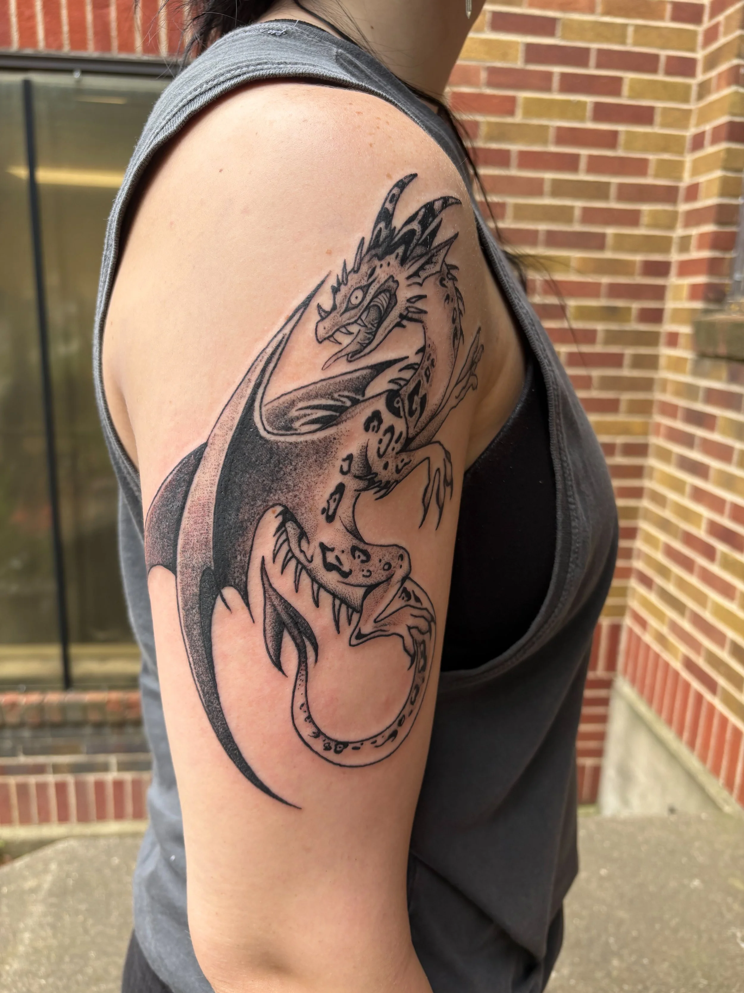 Tattoo of a dragon on woman's upper arm, featuring detailed scales, claws, and sharp features, in black ink.