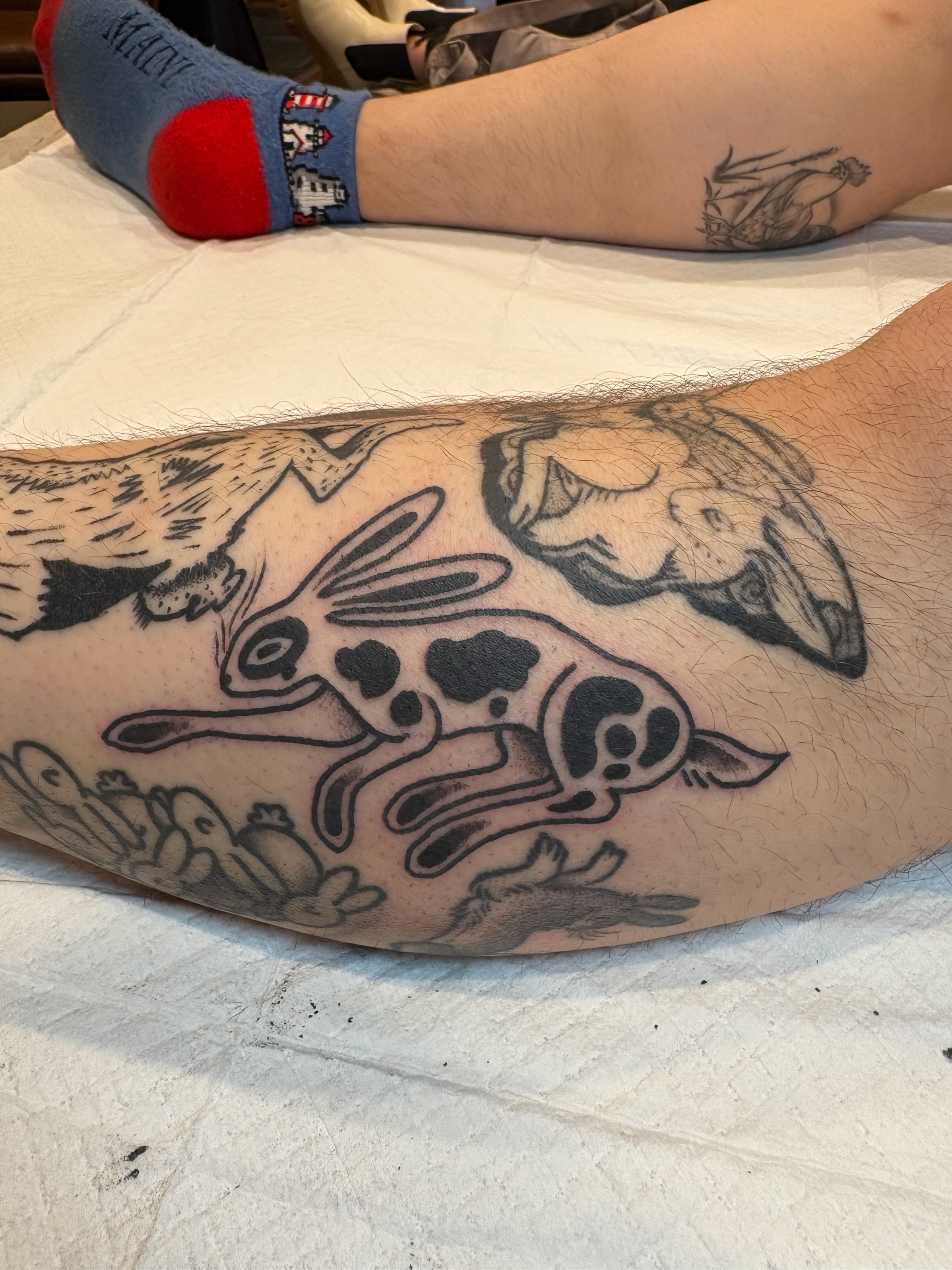 Tattoo on person's leg featuring a black outline of a hare with exaggerated features and spots, surrounded by other tattoos of animals and floral designs.