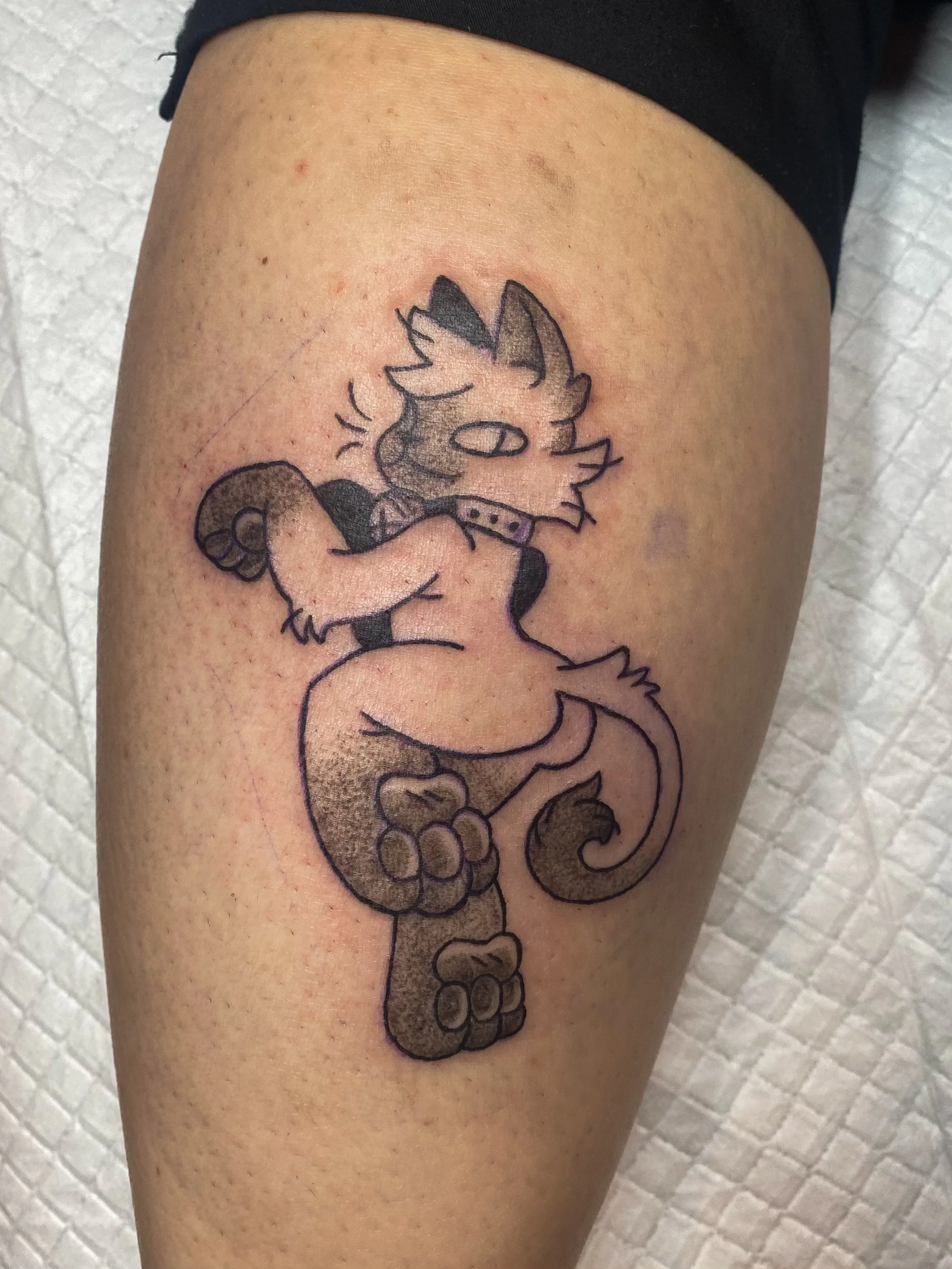 Tattoo of a cartoonish dog character with a long tail and collar, located on a person's upper thigh.