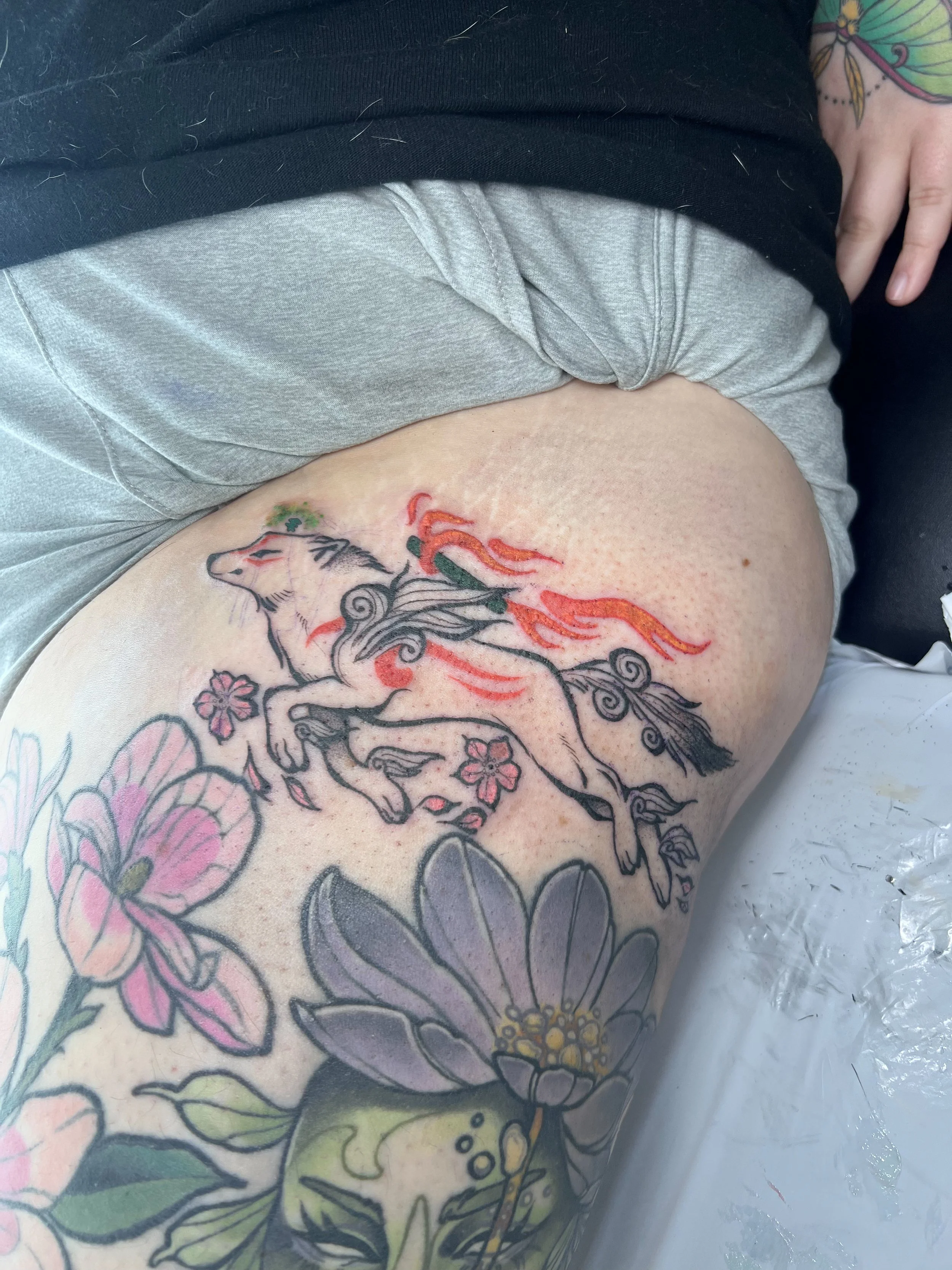 Tattoo of a galloping unicorn with a flowing mane and tail, surrounded by pink and purple flowers, on a person's thigh.