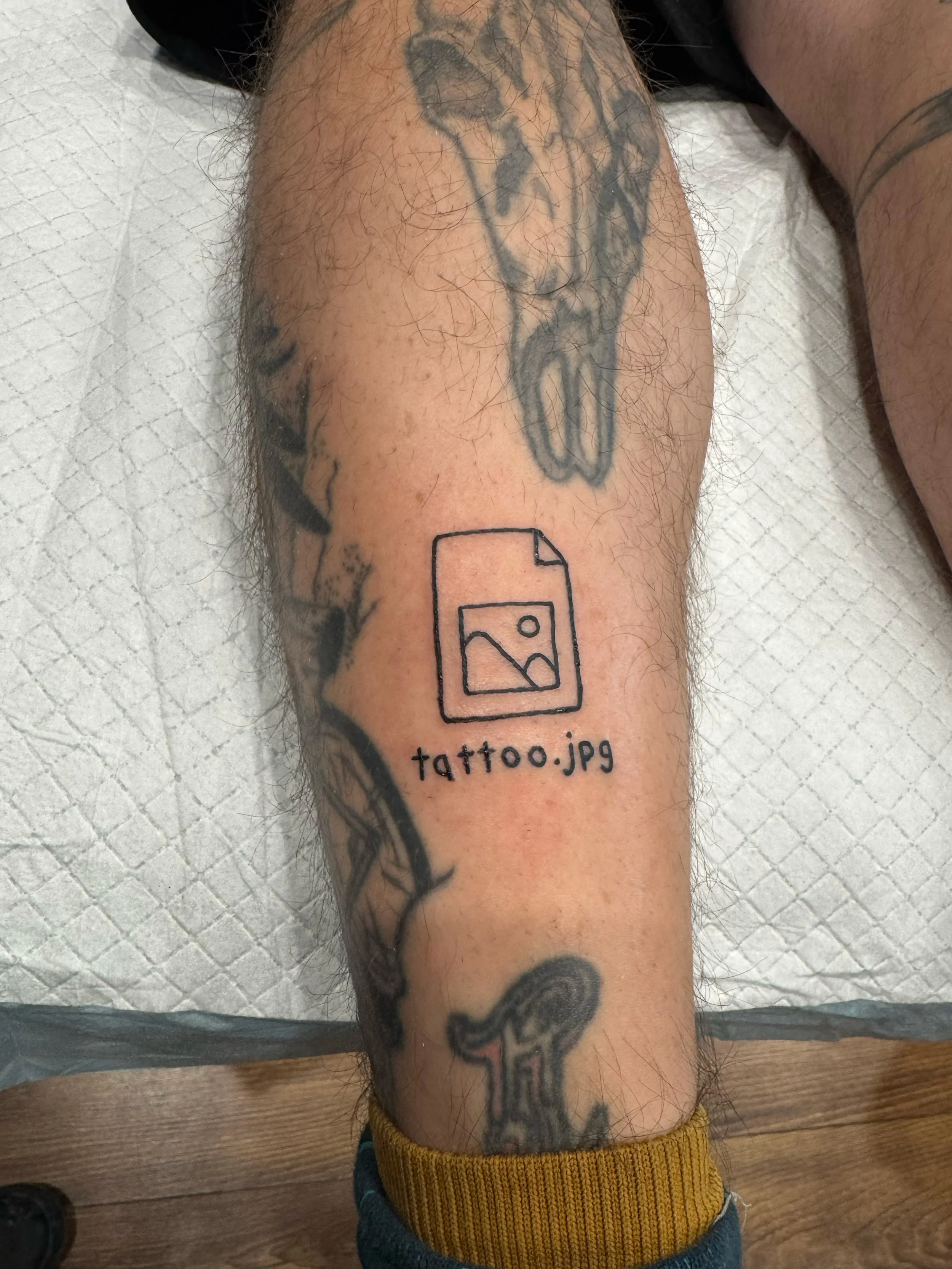 Tattoo of a simple outline of a photo file icon with the text 'gpt.tattoo' underneath, on a person's leg.