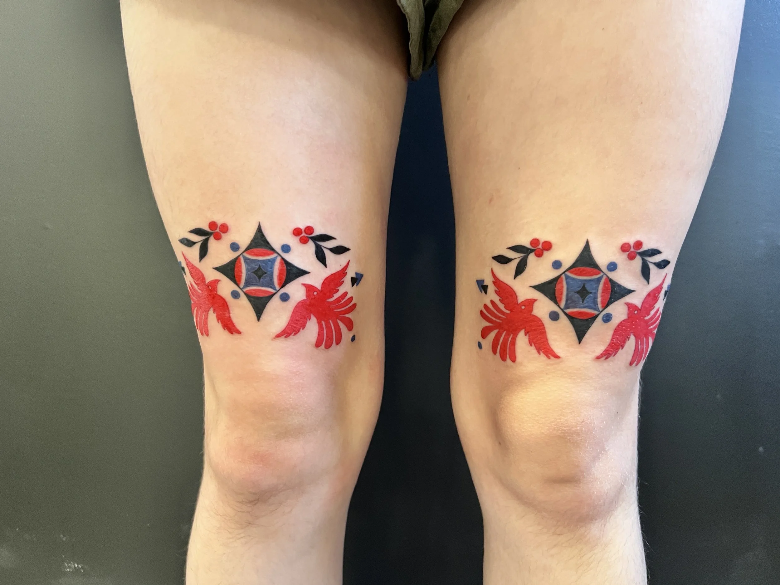 Close-up of a person's thighs with colorful tattoo designs featuring red birds, black leaves, and geometric patterns.