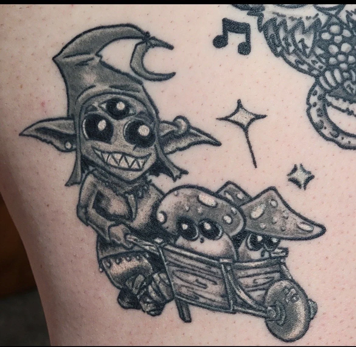 Tattoo of a cartoon goblin riding a wheelbarrow with little skulls and mushrooms, surrounded by stars and musical notes.