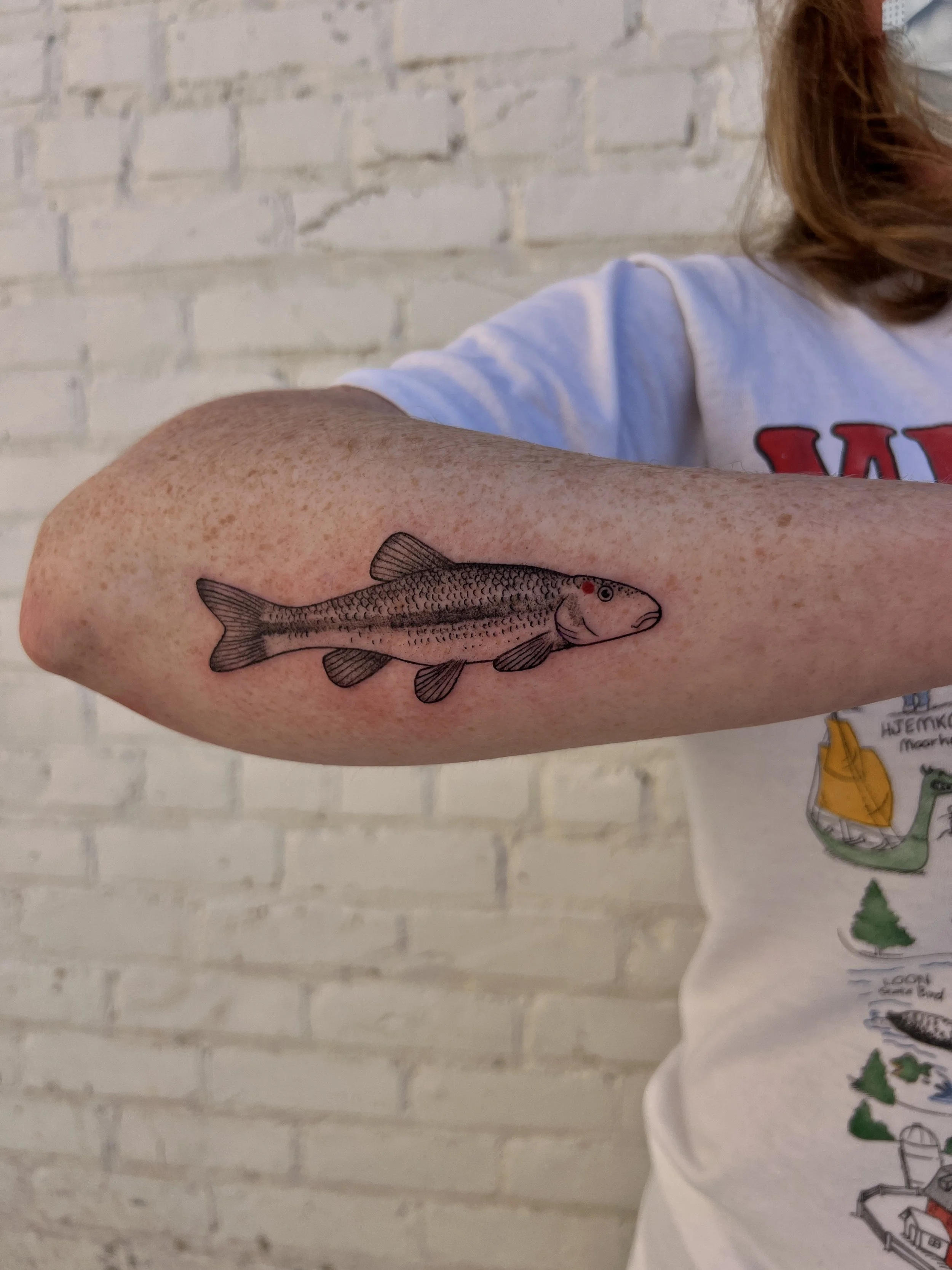 A person with red hair showing a tattoo of a fish on their forearm. The person is wearing a white t-shirt with some colorful drawings and text, and the background is a white brick wall.
