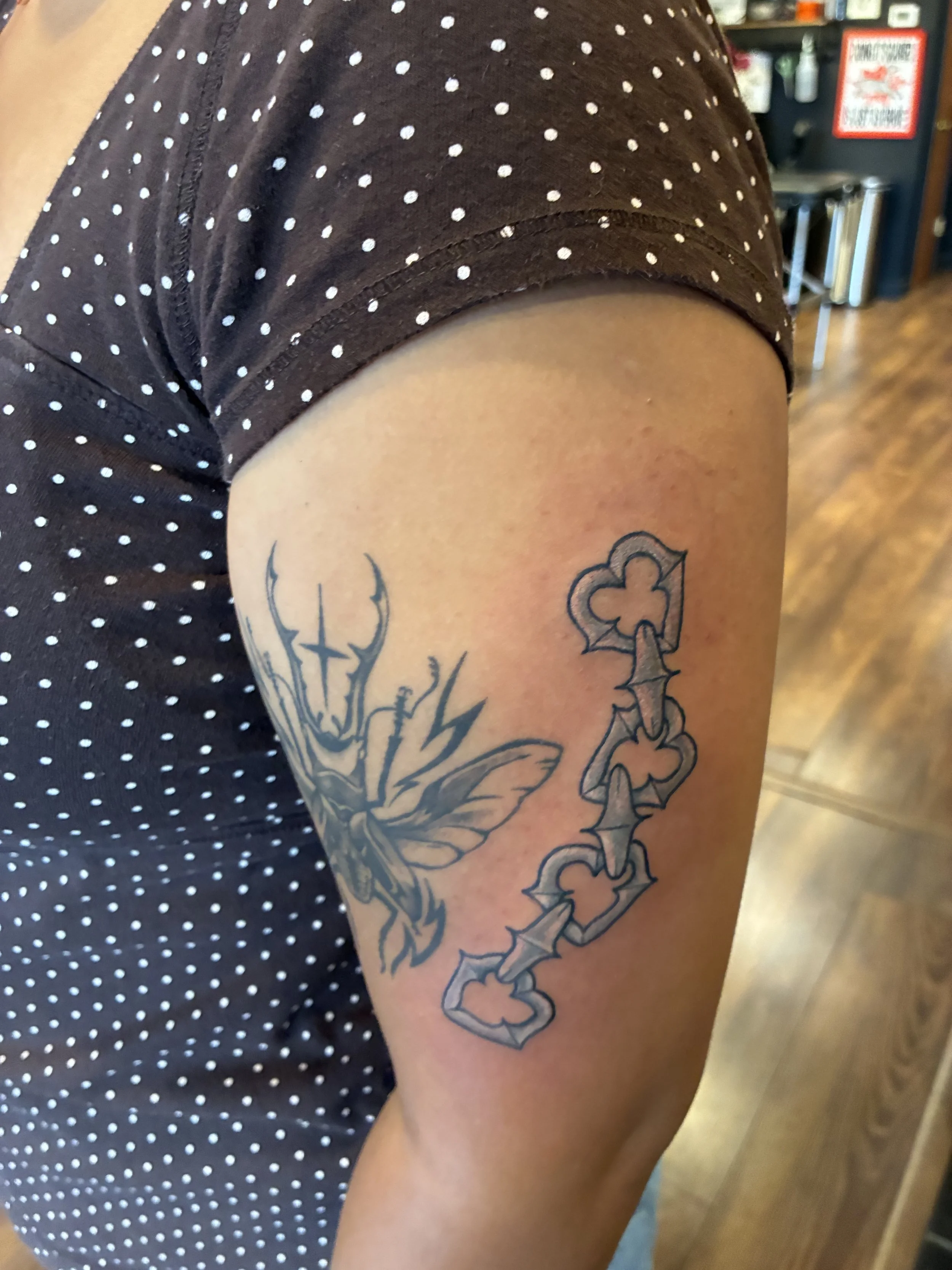 A person's arm with tattoos, including a large flower and a chain of four-leaf clovers, visible next to a black polka-dot shirt. The background includes a wooden floor and a store or cafe setting.