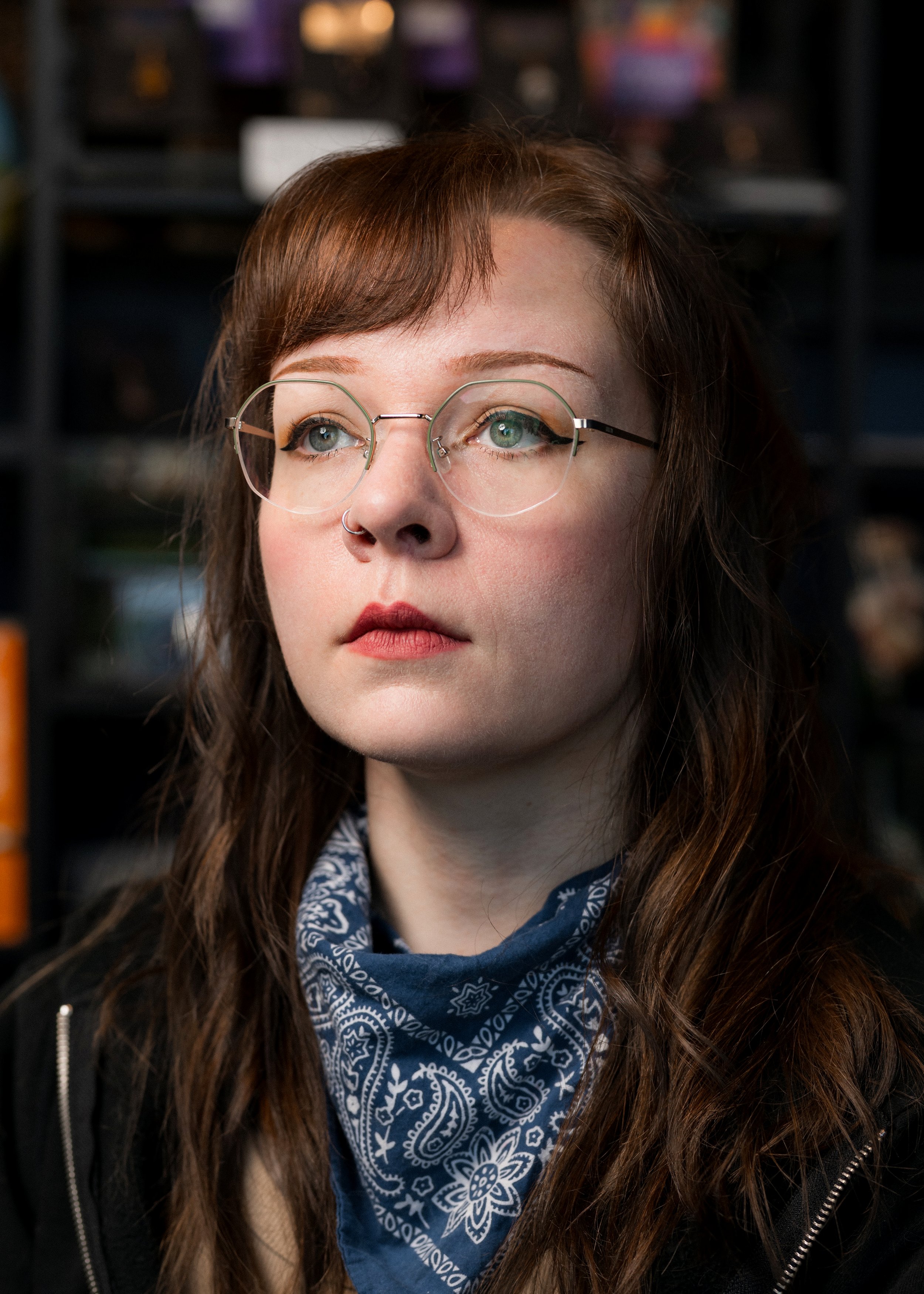 A young woman with long, wavy brown hair and light skin looking to the side. She is wearing round glasses, a nose ring, a blue bandana around her neck, and a black jacket. The background is blurred with shelves and objects.