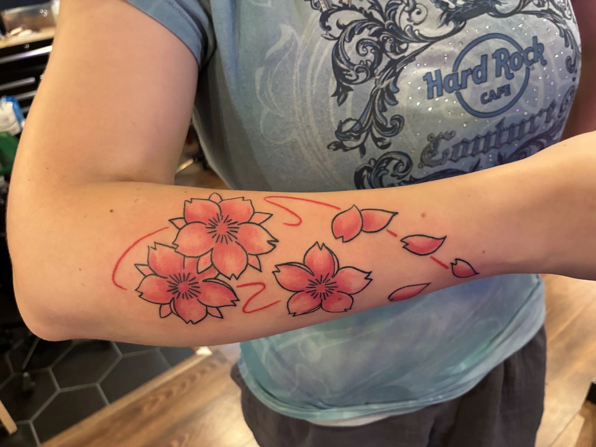 A person wearing a gray Hard Rock Cafe T-shirt with a decorative design. They have a floral tattoo on their forearm with red and black outlines. The background includes a counter and honeycomb floor tiles.