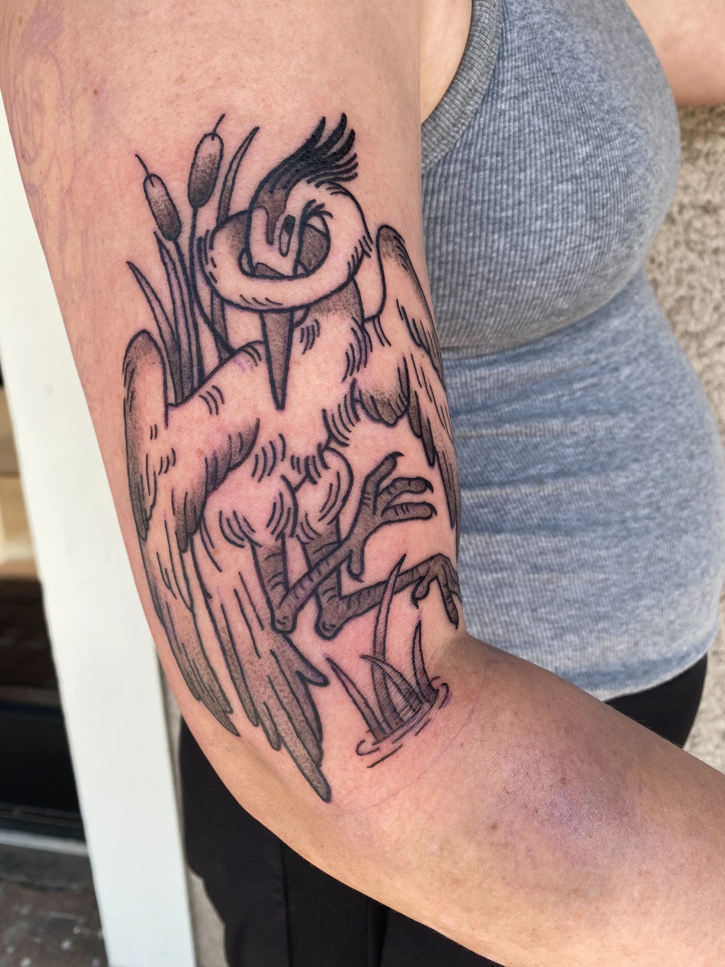 Black outlined tattoo of a bird with a branch in its beak on a person's upper arm, with part of a gray tank top visible.
