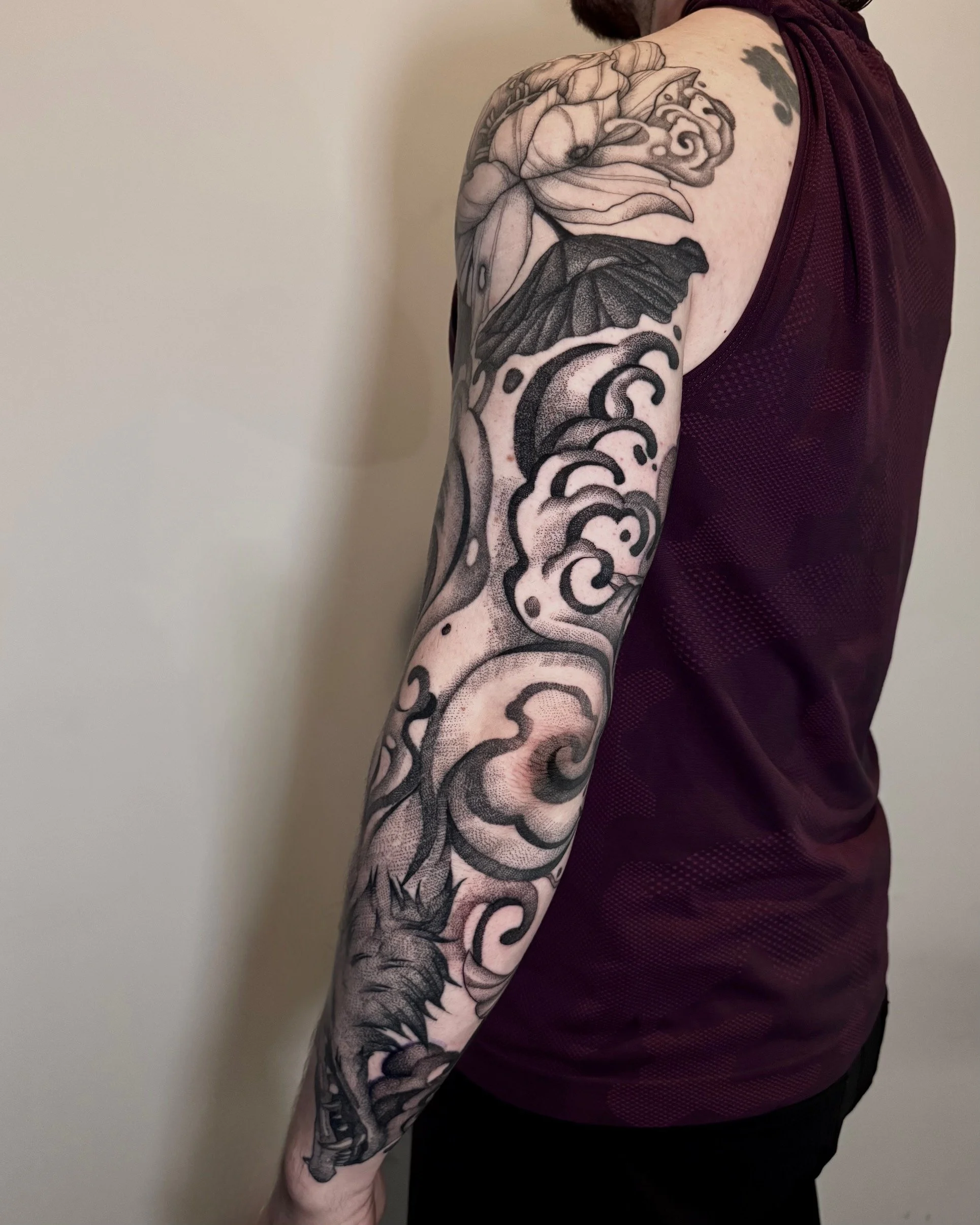 A person's arm with a detailed black and gray tattoo sleeve featuring a dragon, clouds, and floral elements.
