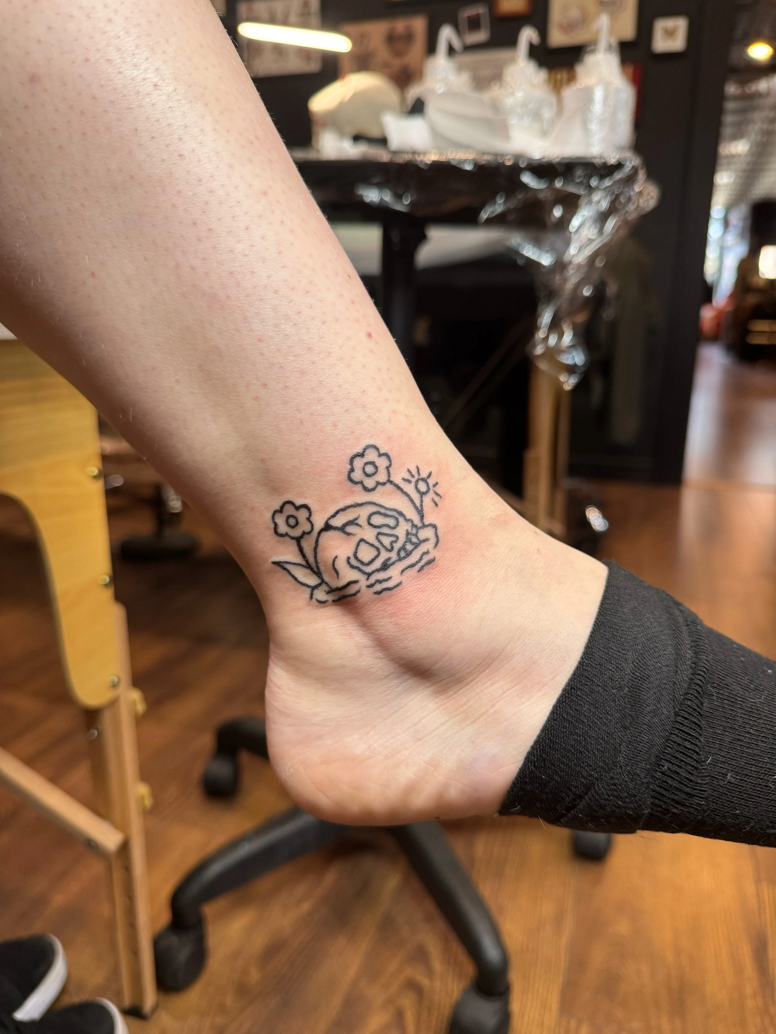 Tattoo of a skull with a flower, plant, and sun on a person's ankle.