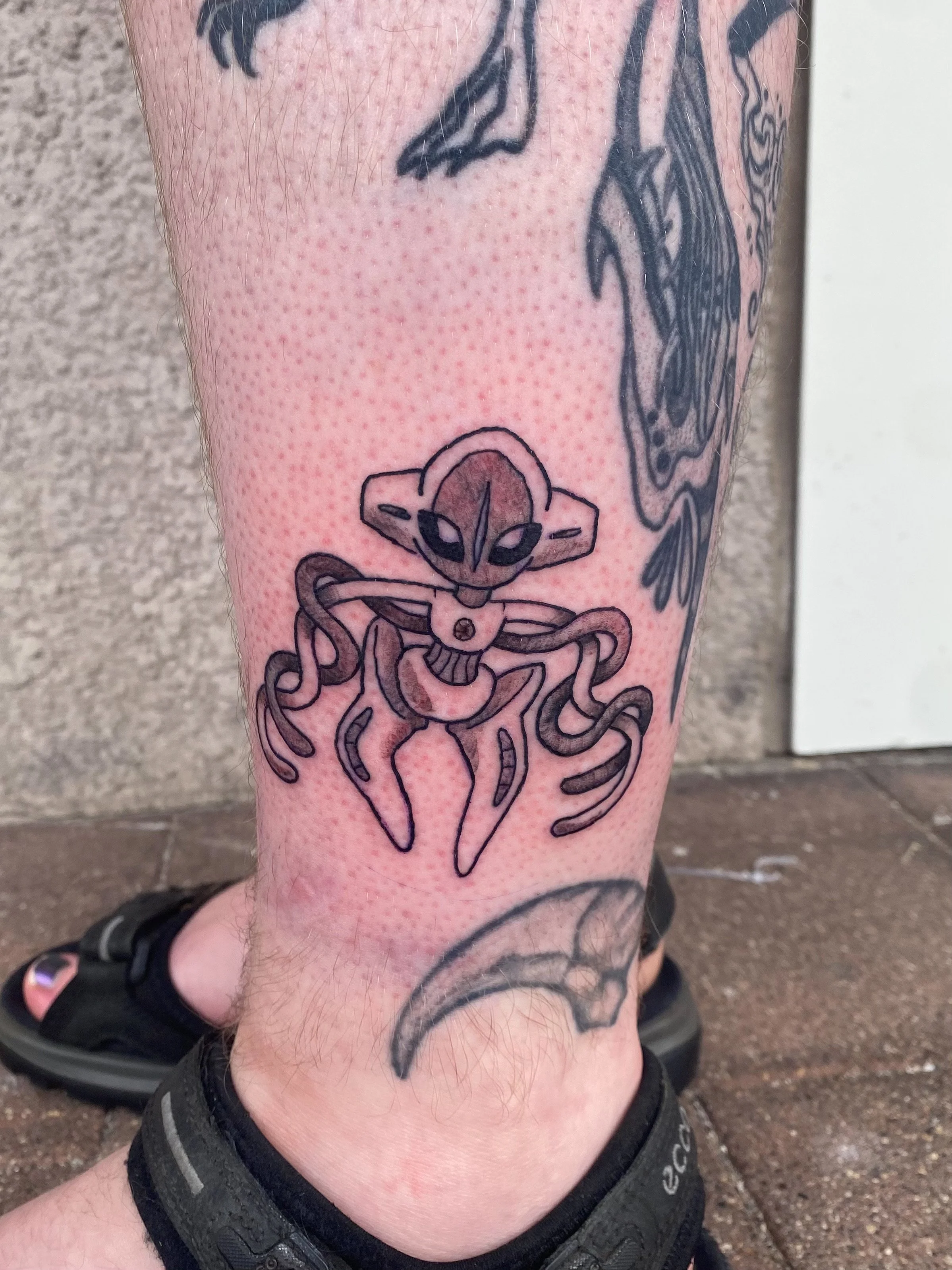Tattoo of a stylized charmander Pokémon on a person's lower leg with a black strap around the ankle, standing on a tiled surface.