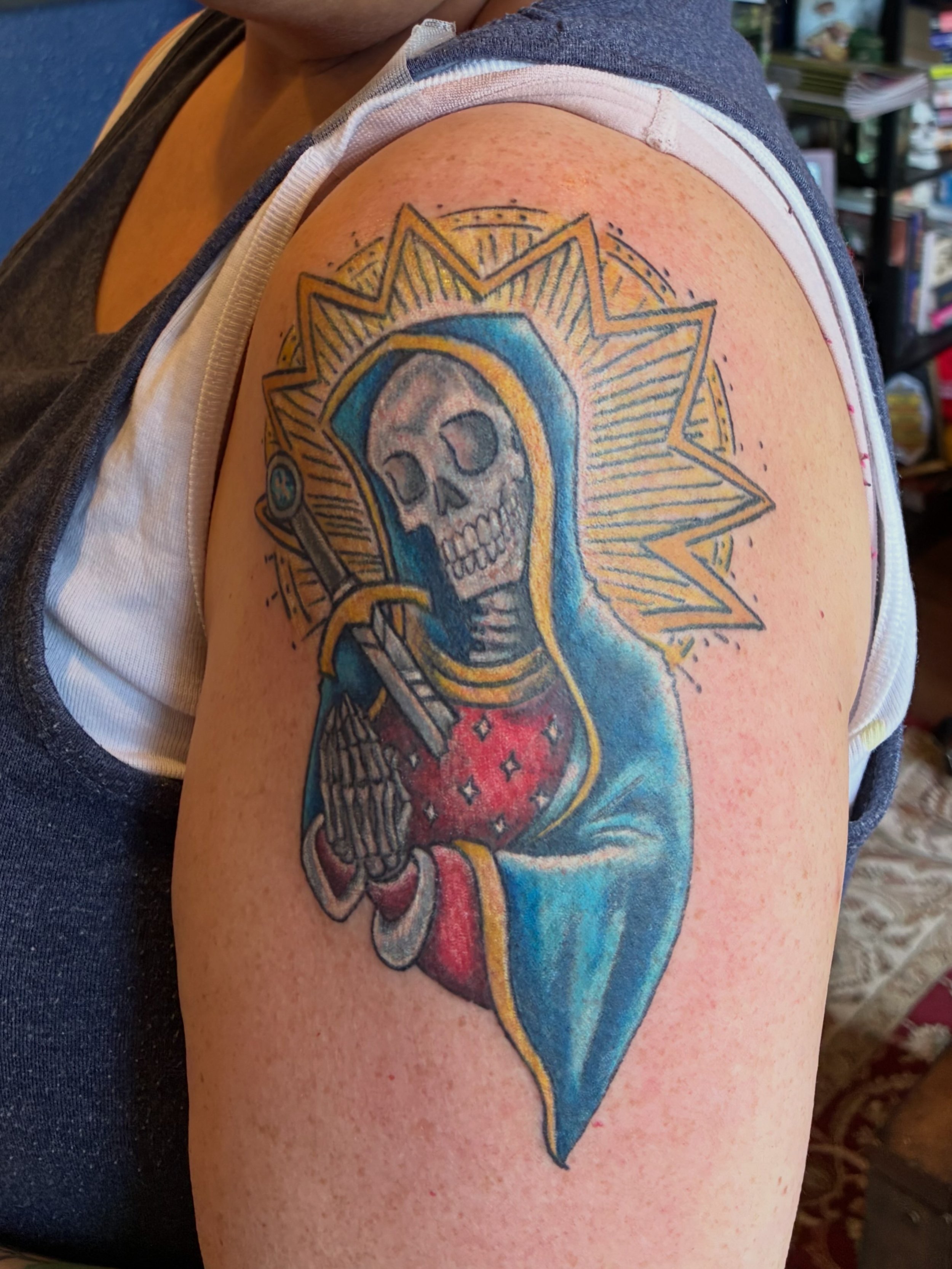 Colorful tattoo of a skeleton inside a blue diamond shape, with yellow accents and black outlines, on a person's upper arm.