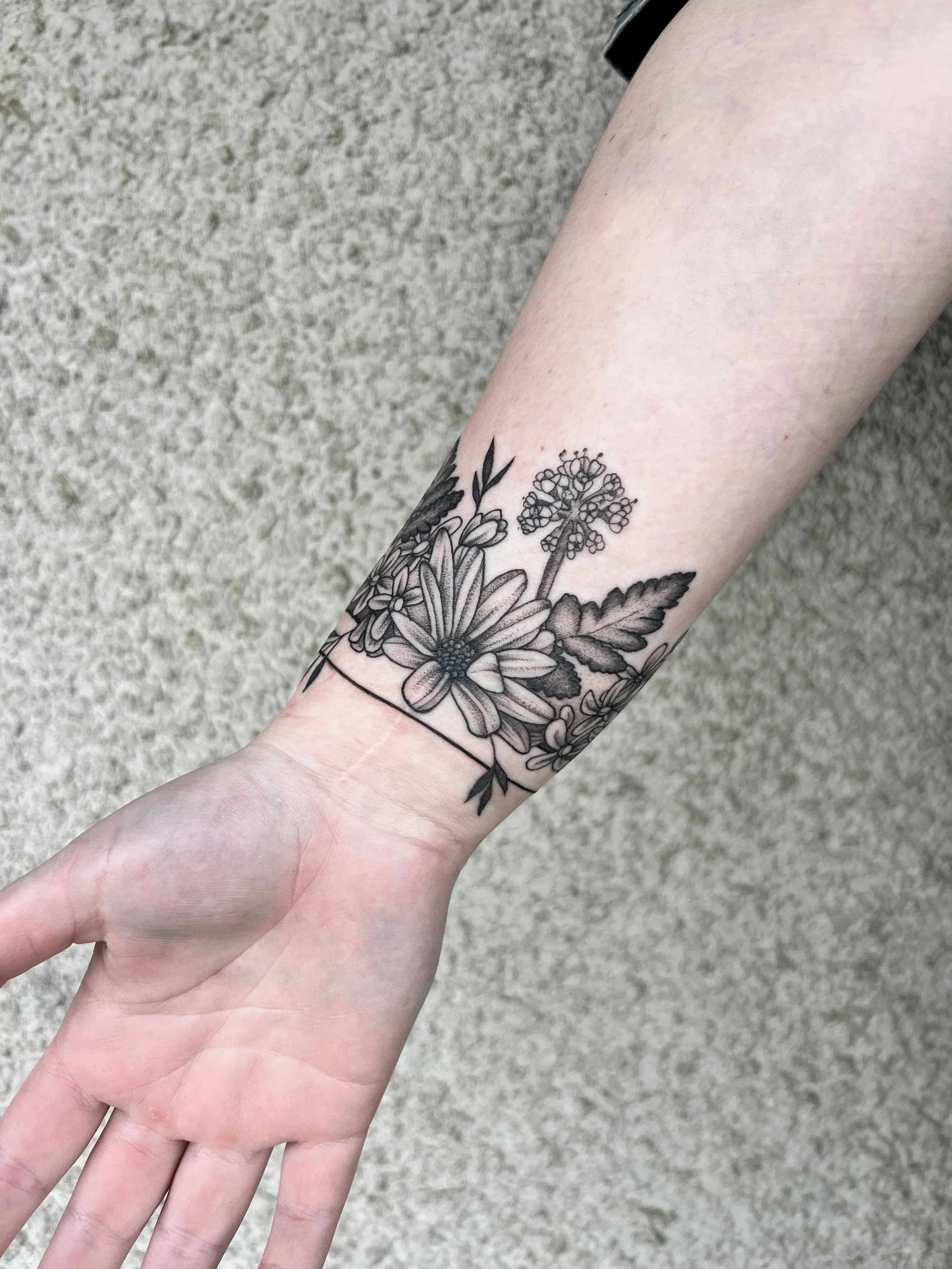 A person's arm with a black floral tattoo, showing large daisy-like flowers, leaves, and small buds, against a neutral textured background.