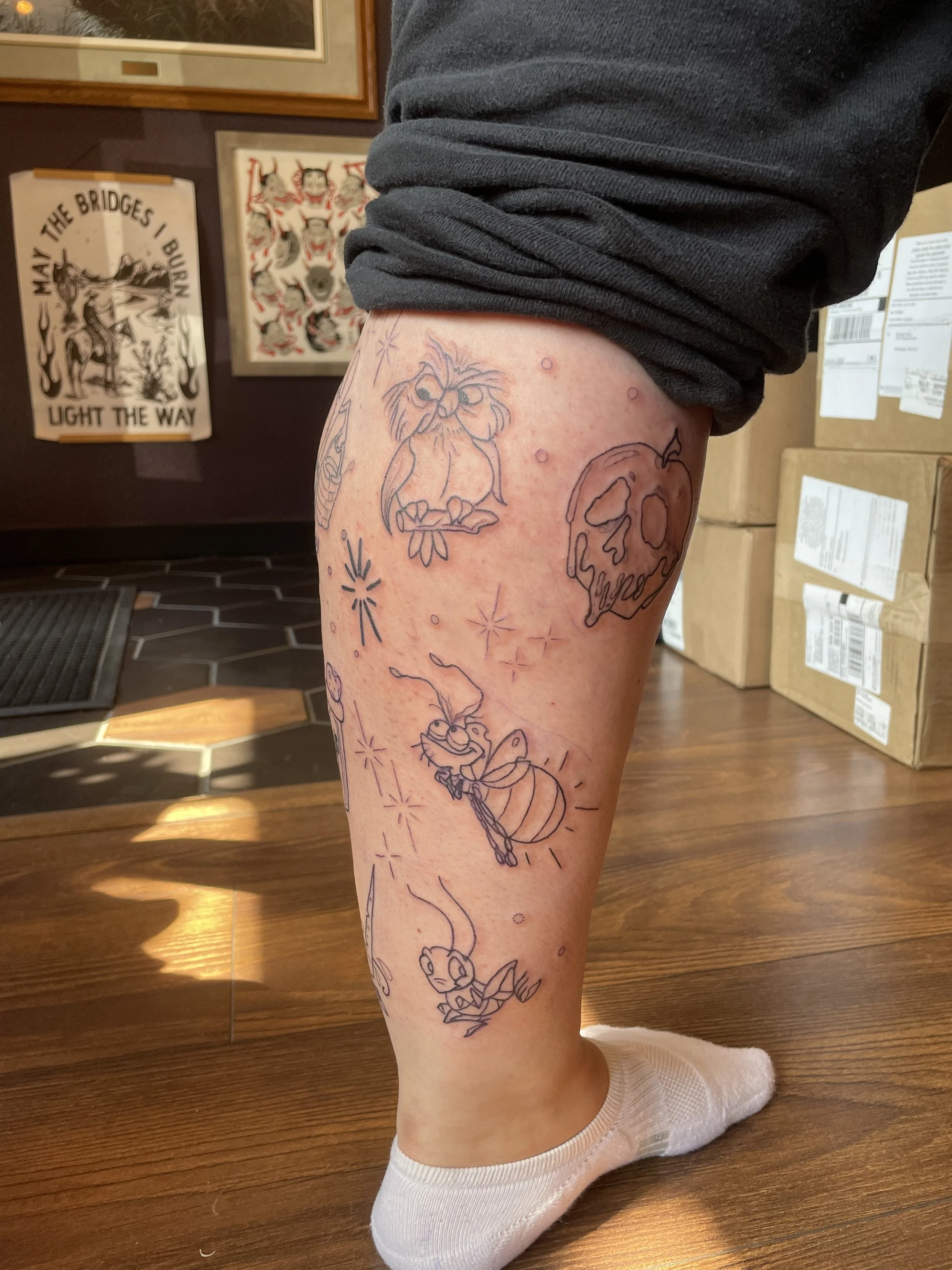 Unfinished tattoo designs on a person's thigh, including characters and objects from the movie 'Sesame Street' such as Grover, a bee, and a skull-shaped pumpkin, with some outlines and shading yet to be completed.