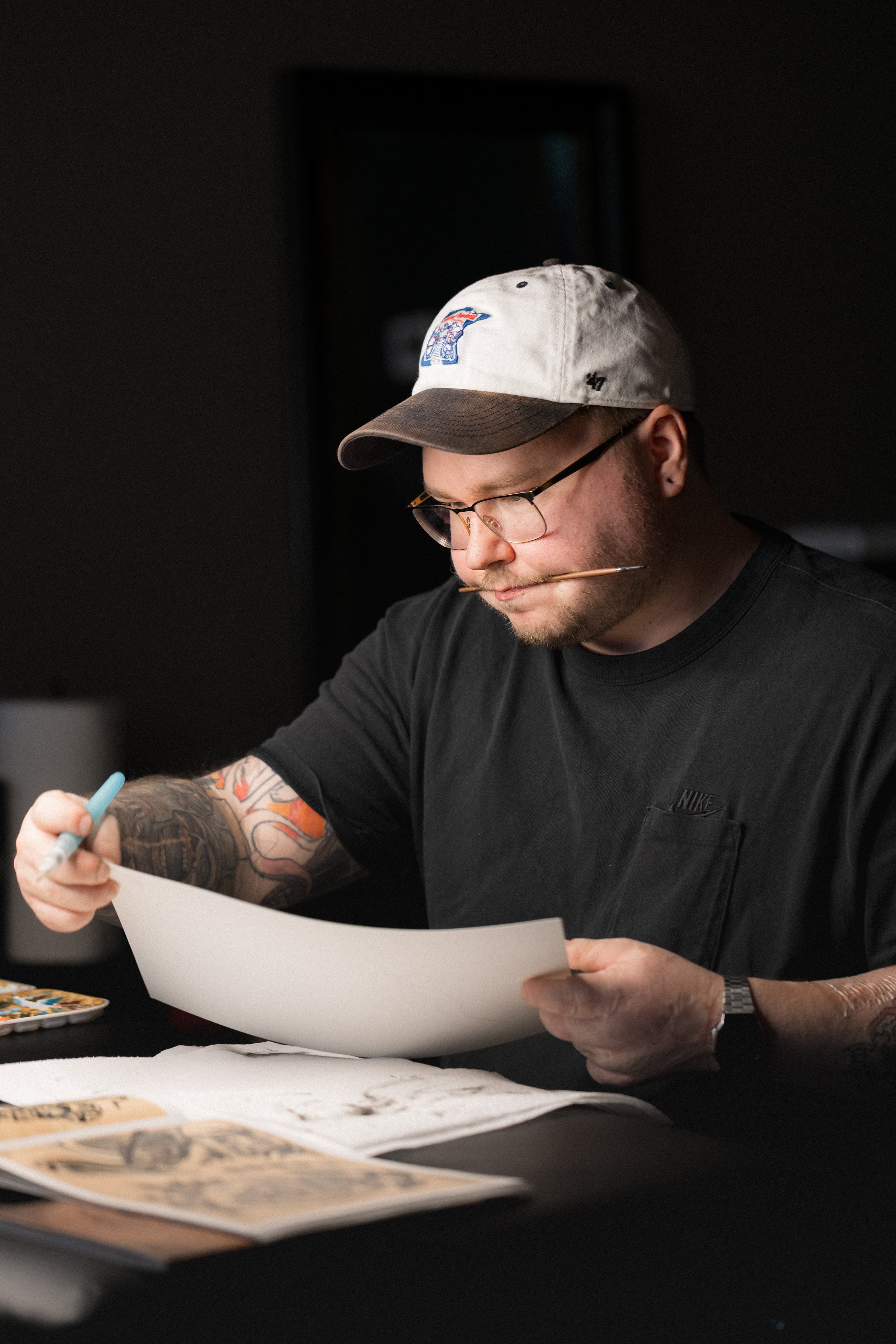 A man with glasses, tattoos, and a hat is sitting at a table, studying papers and illustrations.