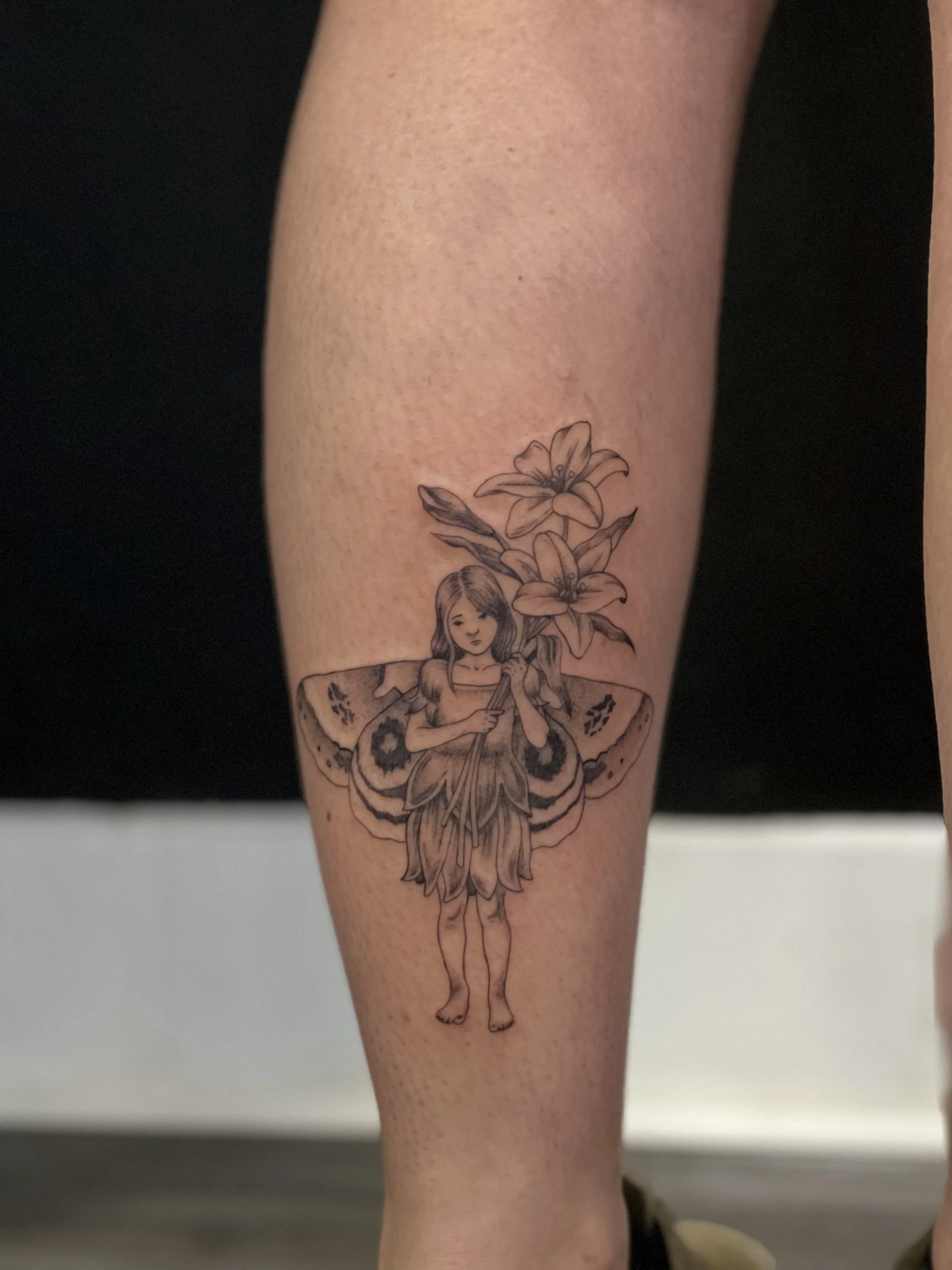 Black and white tattoo on person's arm of a fairy with large butterfly wings, flowers, and a girl holding a sword in the center.