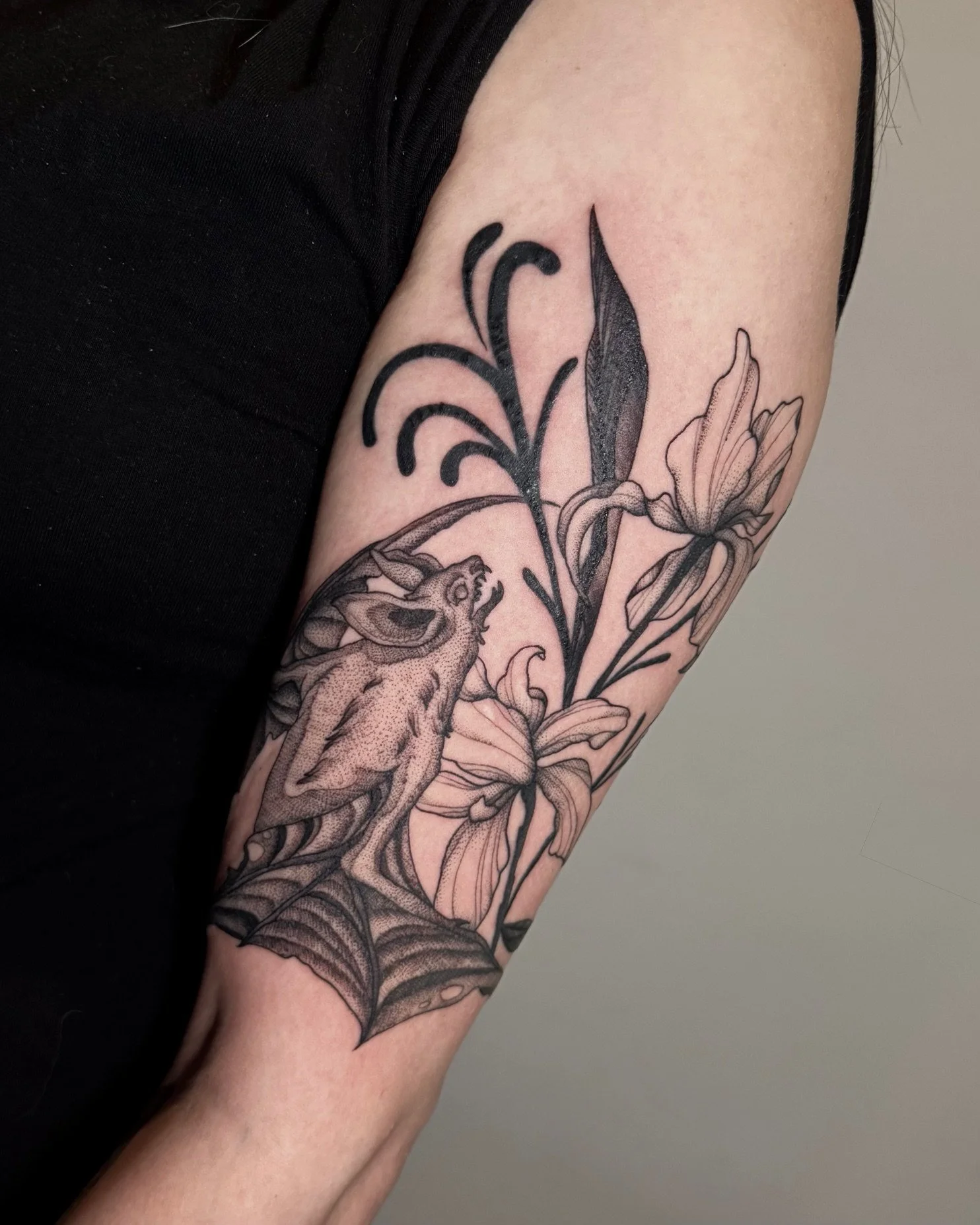 Black and gray tattoo of a bat and lilies on a person's upper arm.
