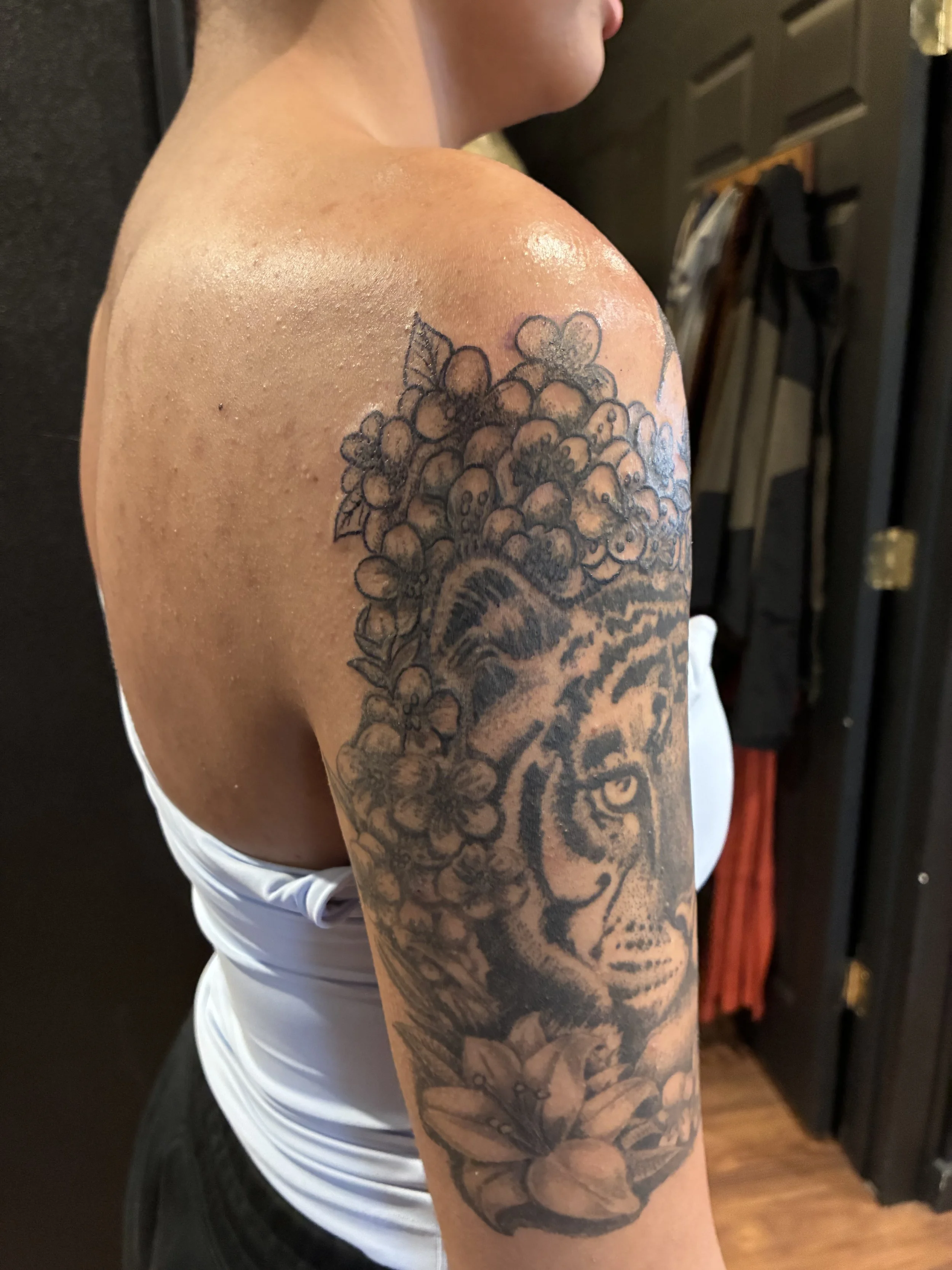 A person's upper arm and shoulder with a black floral tattoo, including flowers and leaves, against a background of a dark room and wardrobe.