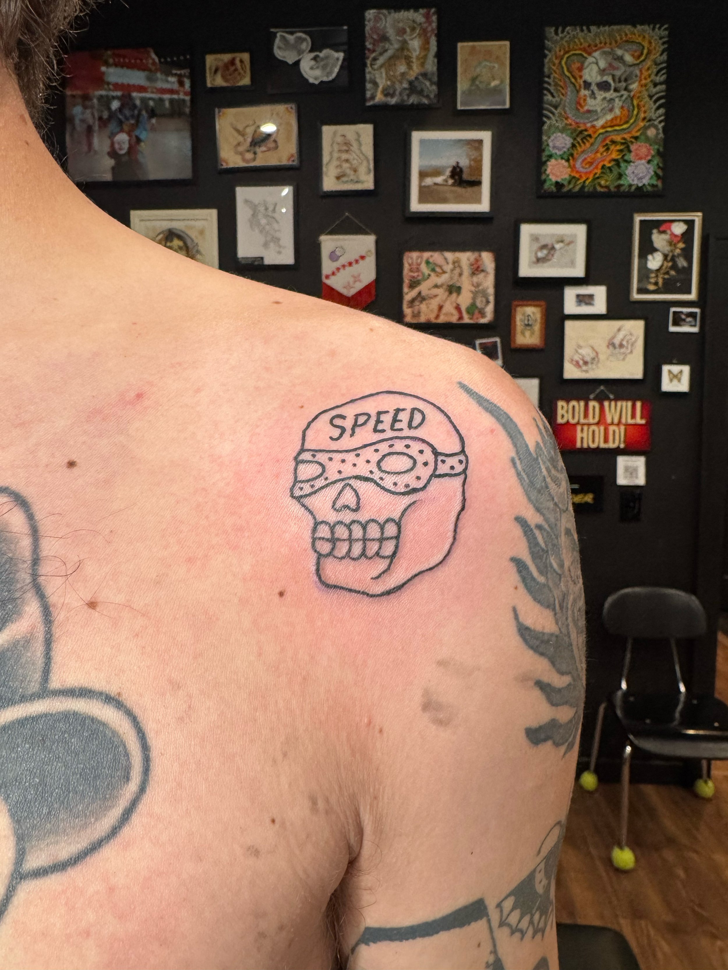 Tattoo of a skull with the word 'SPEED' on the forehead, wearing a racing helmet, on a person's shoulder.