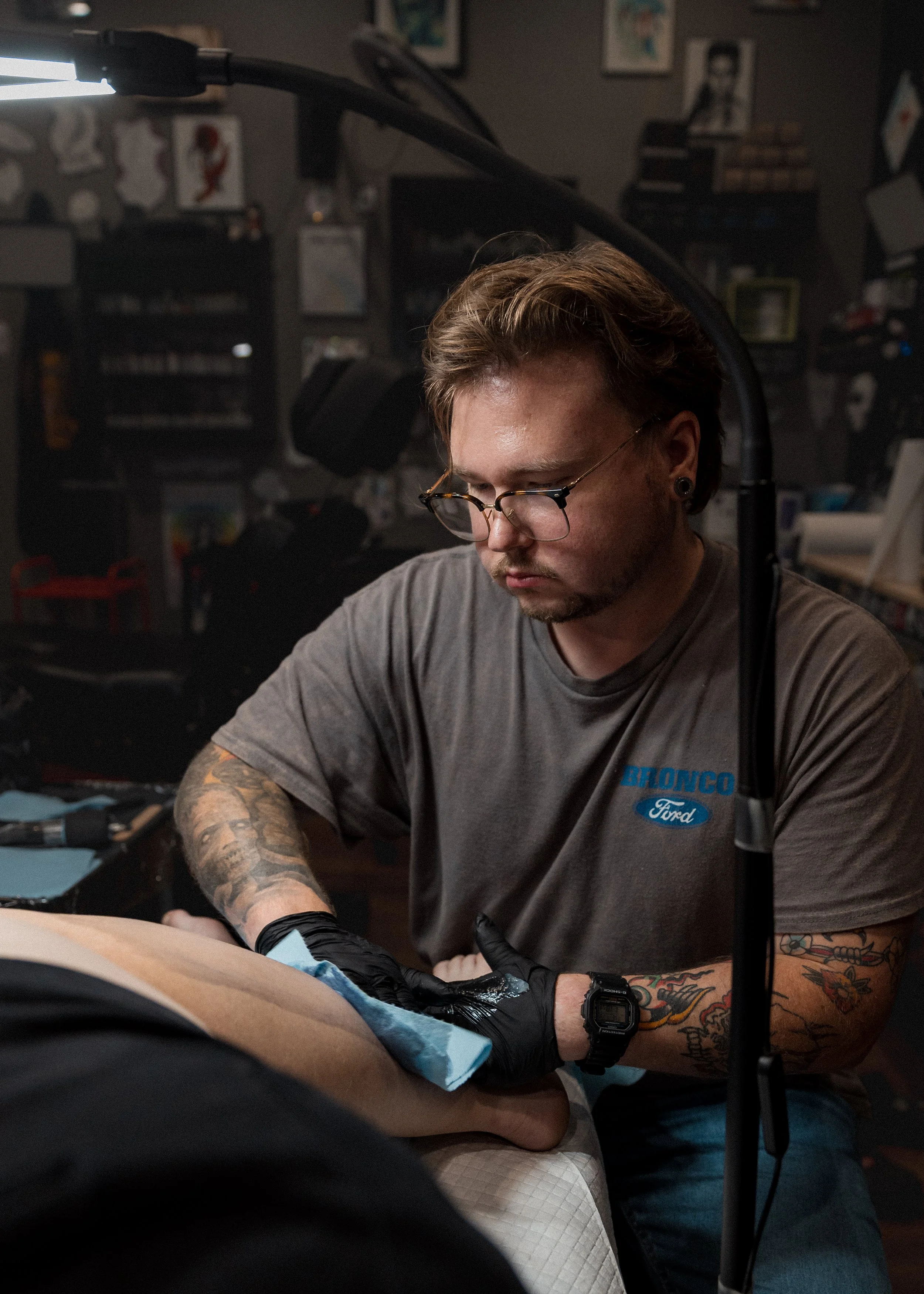 A person with glasses, tattoos, and a Ford Bronco shirt is tattooing a person lying down, in a dimly lit tattoo studio.
