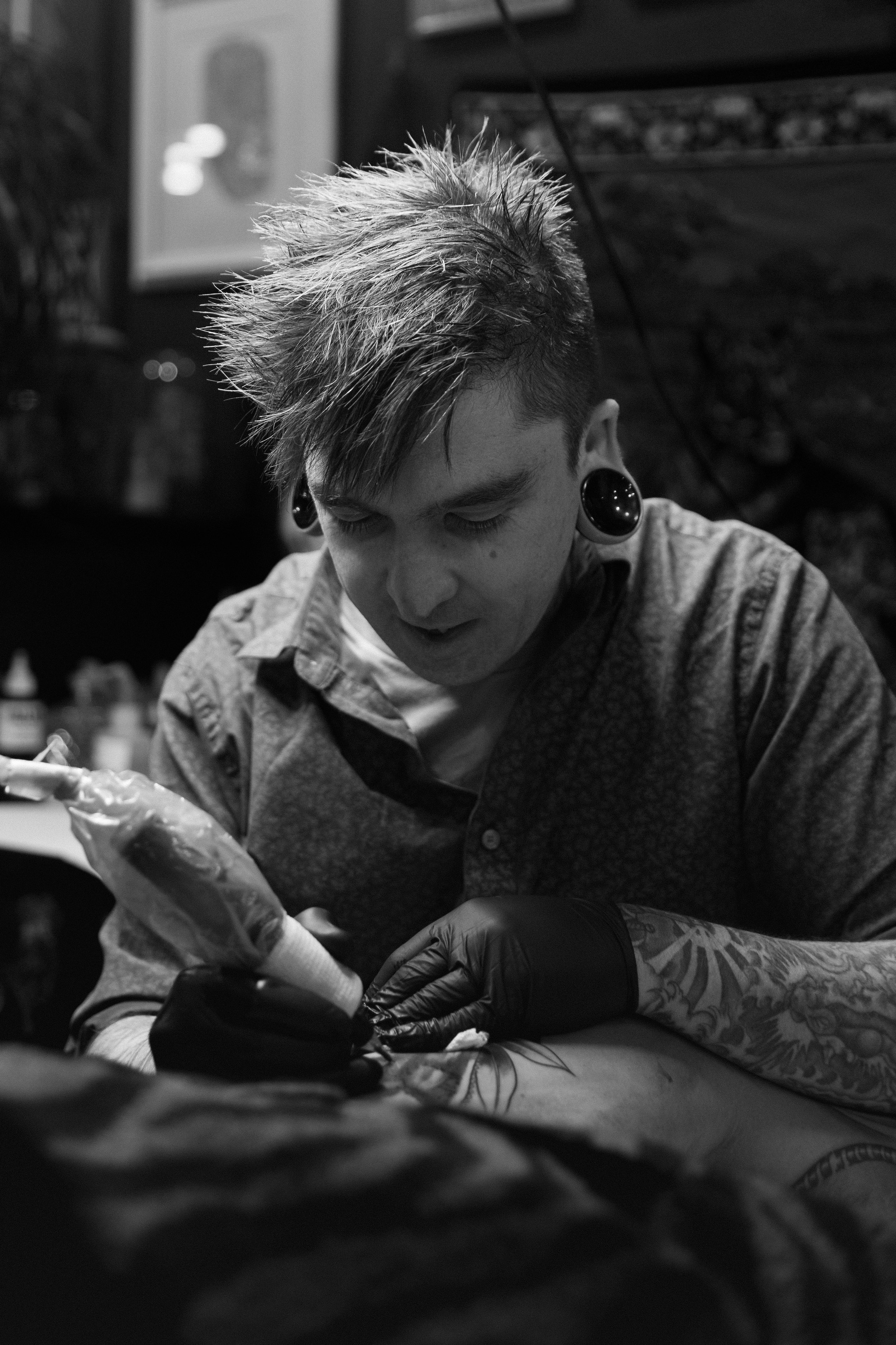 A person with dyed spiky hair and stretched earlobes getting a tattoo on their arm in a tattoo studio.