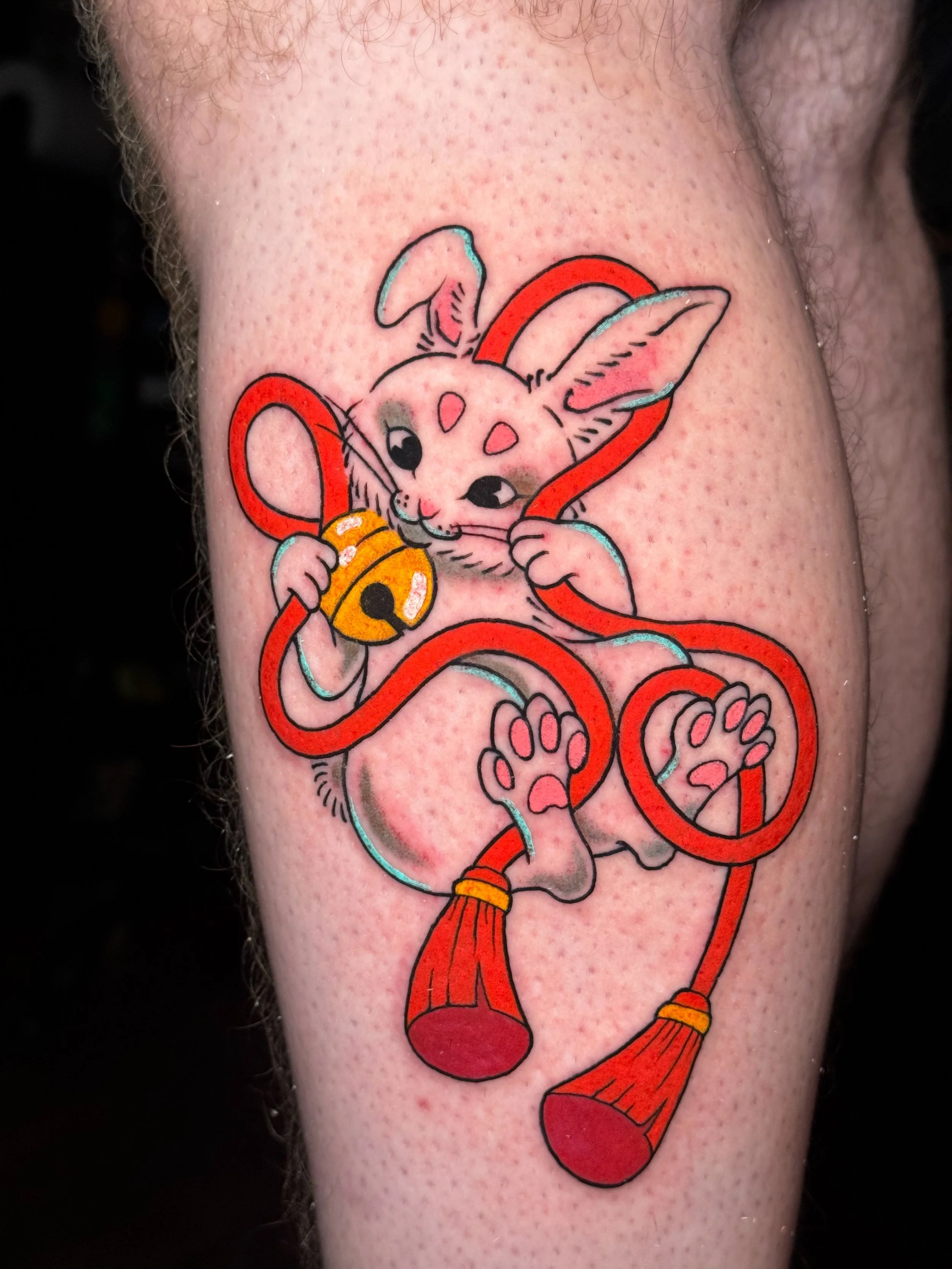 Tattoo of a cartoon bunny holding a yellow bell and wearing a red tie, with red and blue ribbons.