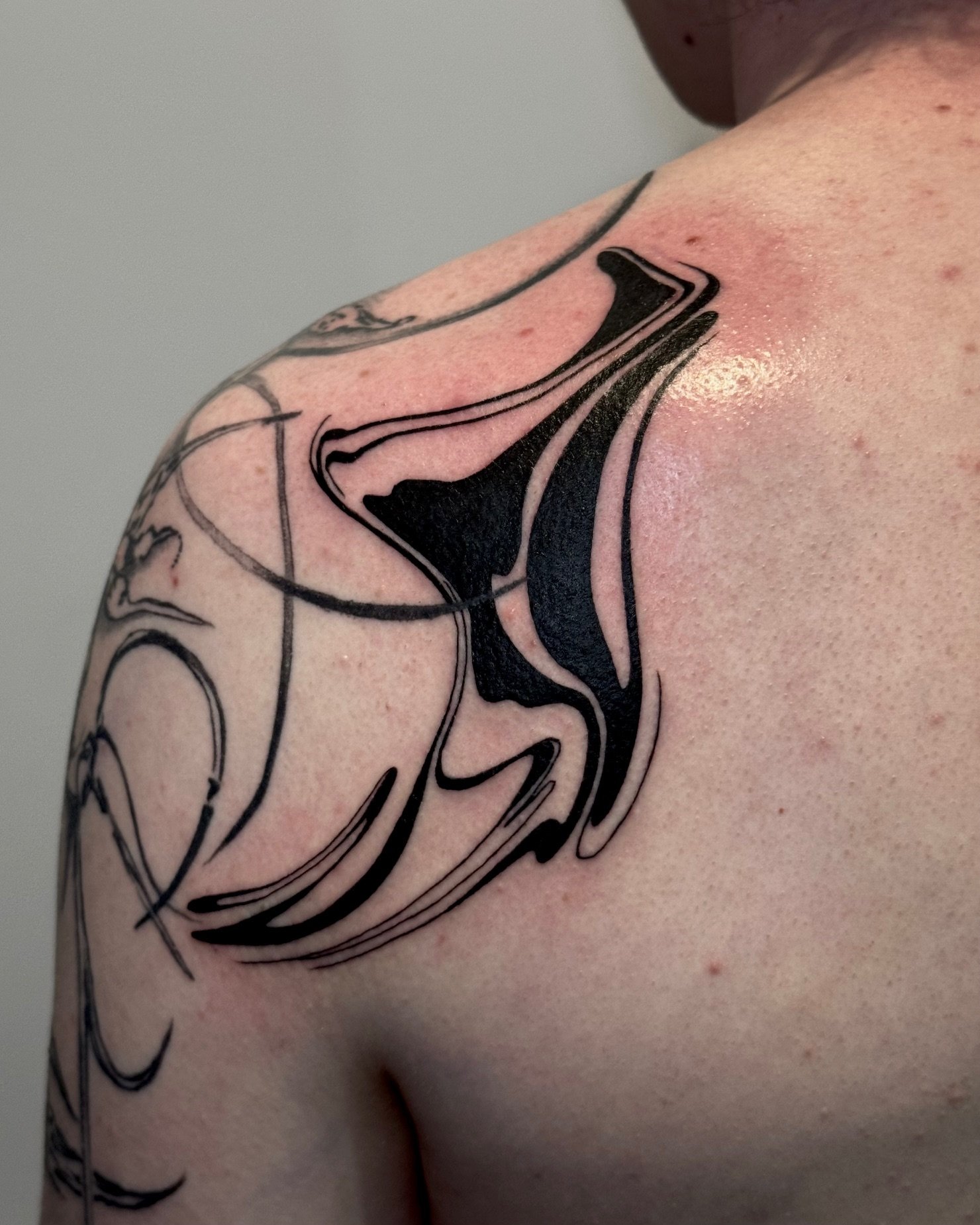 A close-up of a person's shoulder with a black and gray abstract tattoo design.