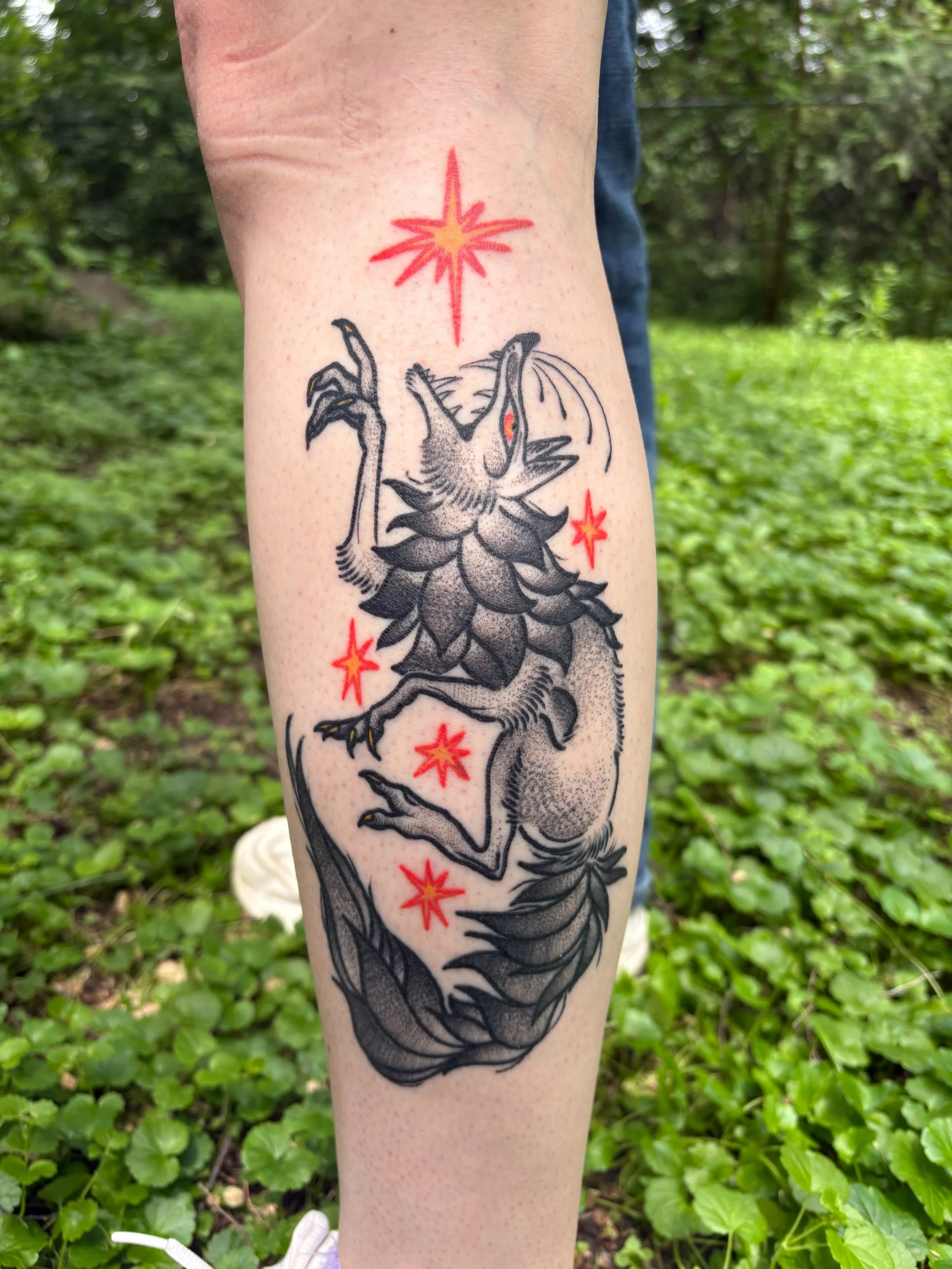 Tattoo of a mythical serpentine creature with a triangulated face, clawed limbs, and an elongated body, surrounded by red and orange starburst designs, on a person's forearm.