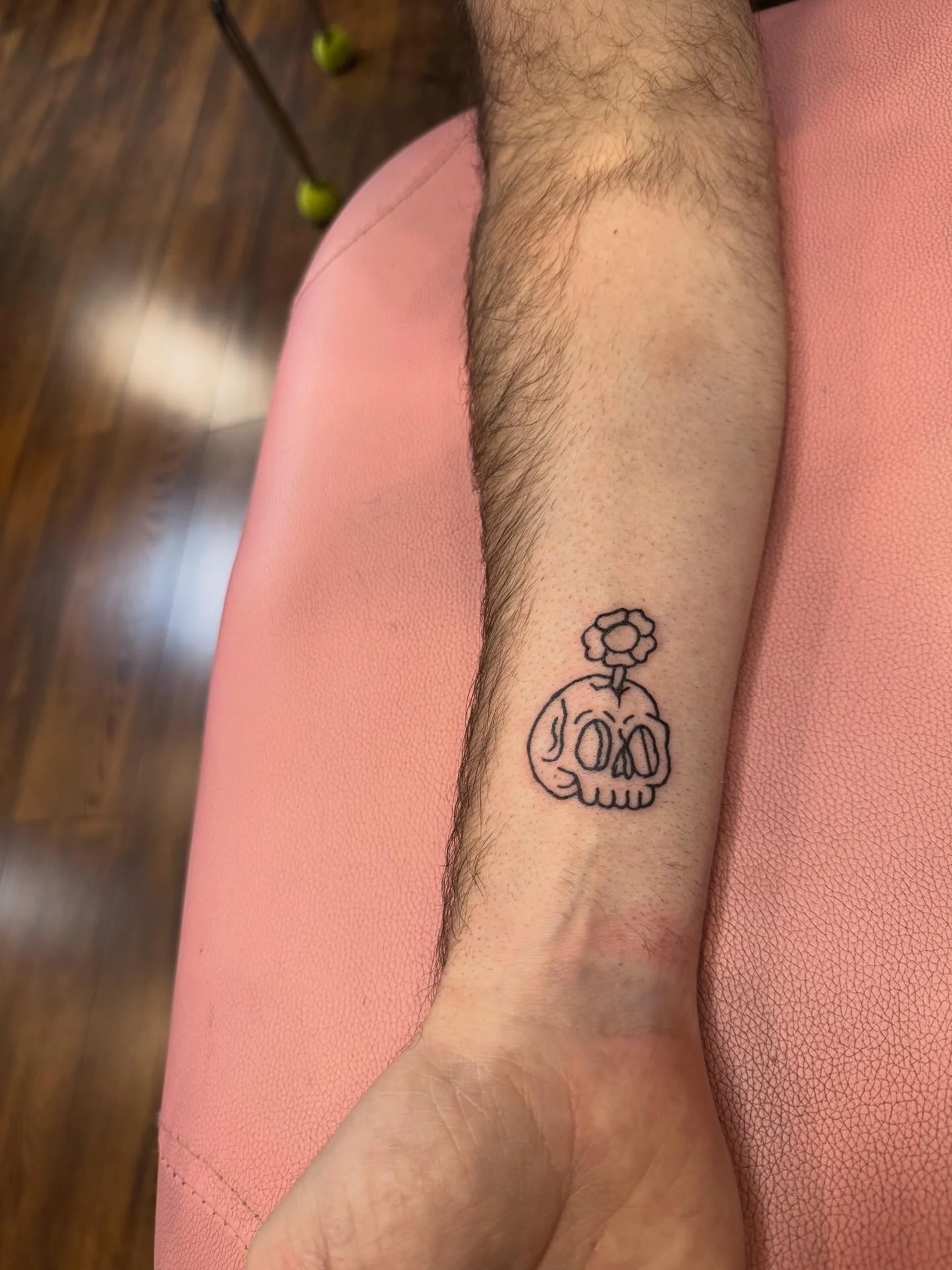 A person's forearm with a tattoo of a pumpkin skull with a flower growing out of the top, drawn with black outline.