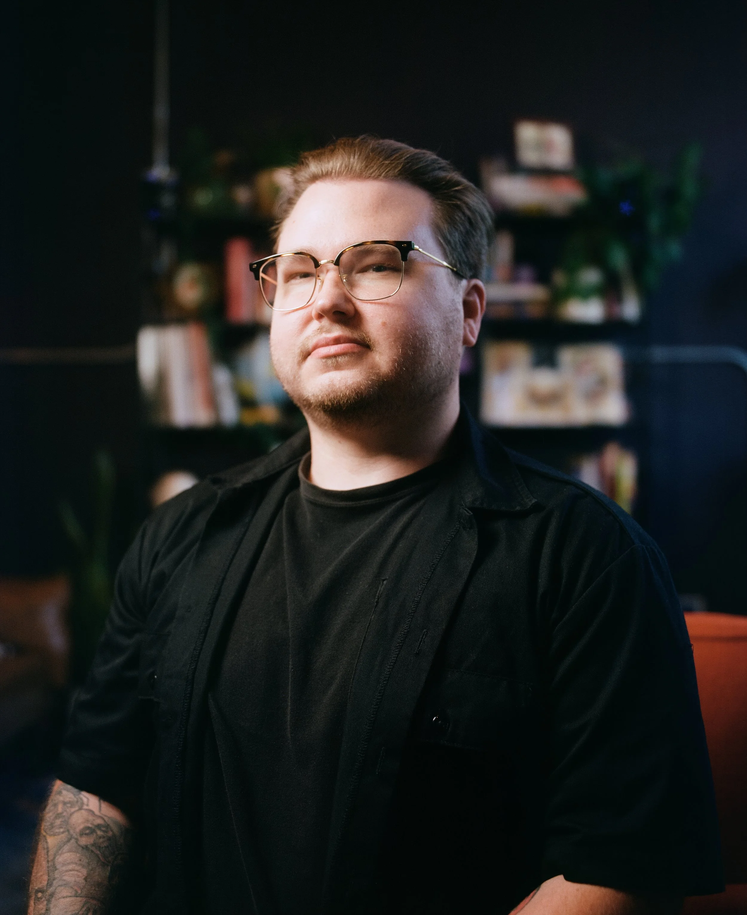 Portrait of a man with glasses and a beard, wearing a black shirt, sitting indoors with a blurred background of books and plants.