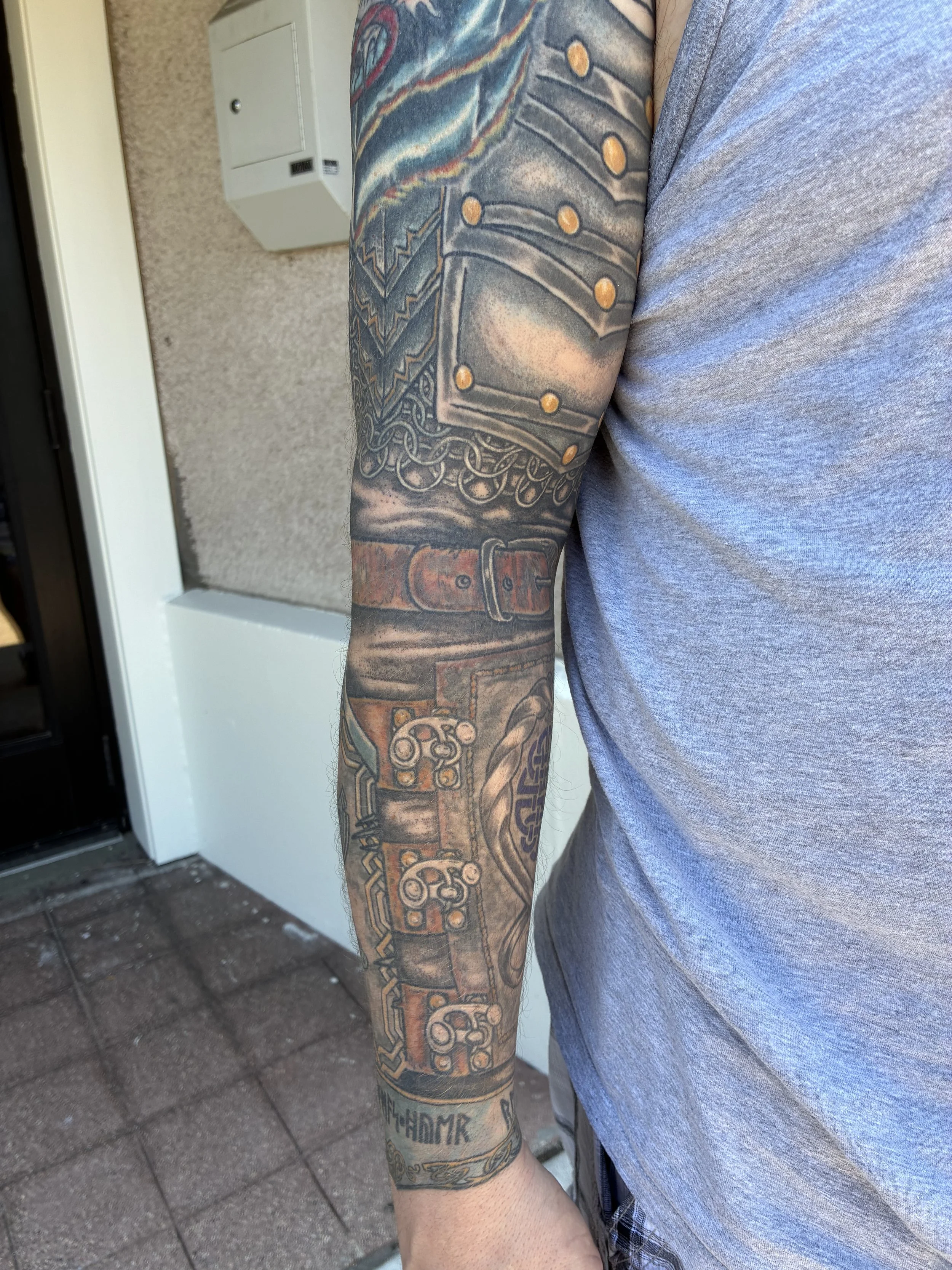 Close-up of a person's arm with colorful tattoo art depicting a gladiator helmet, chains, and intricate designs with a belt tattoo at the wrist, wearing a gray t-shirt.