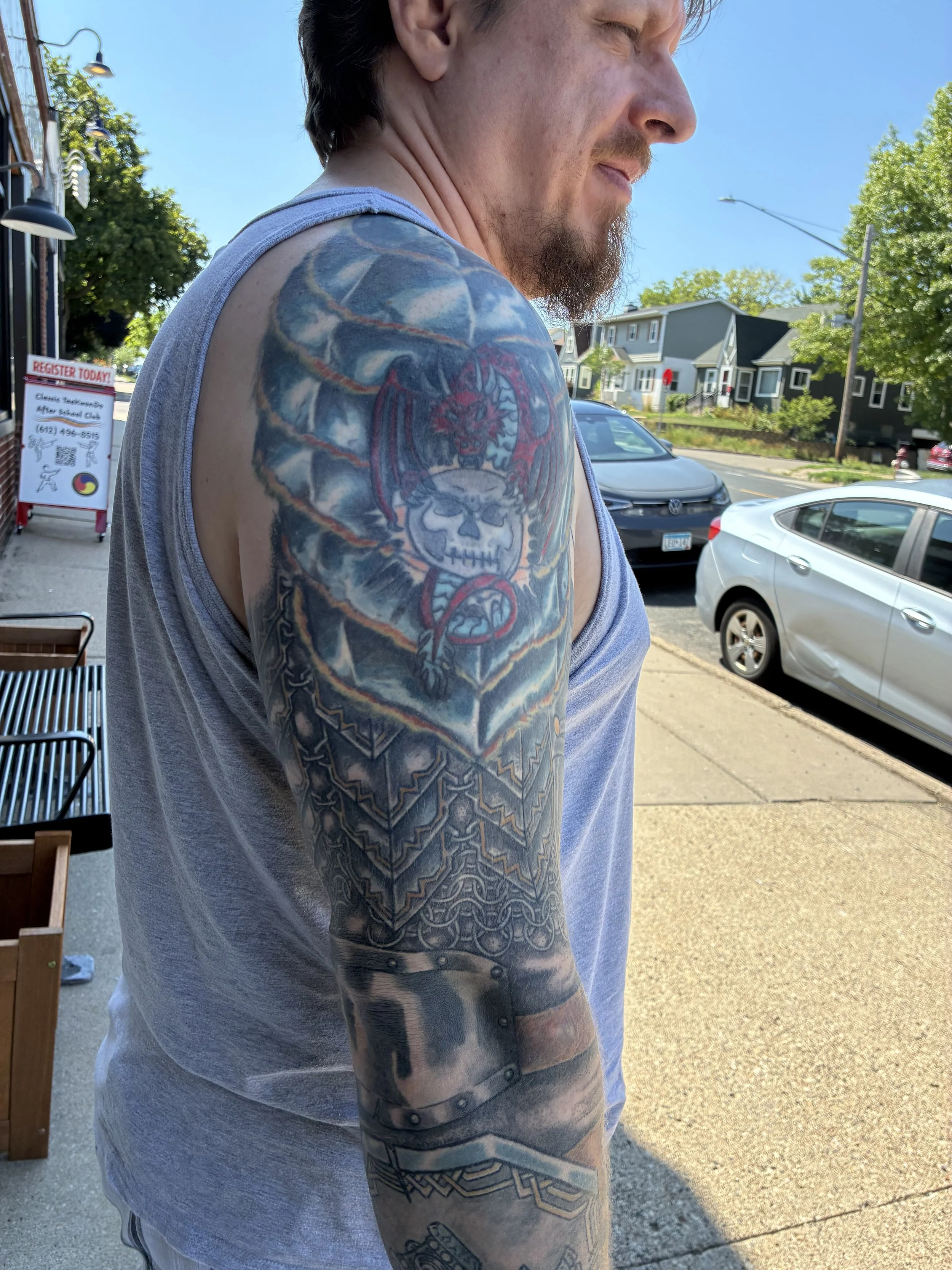 A close-up of a man with a tattoo sleeve on his left arm, standing outdoors on a sunny day, wearing a light sleeveless shirt, with parked cars and houses in the background.