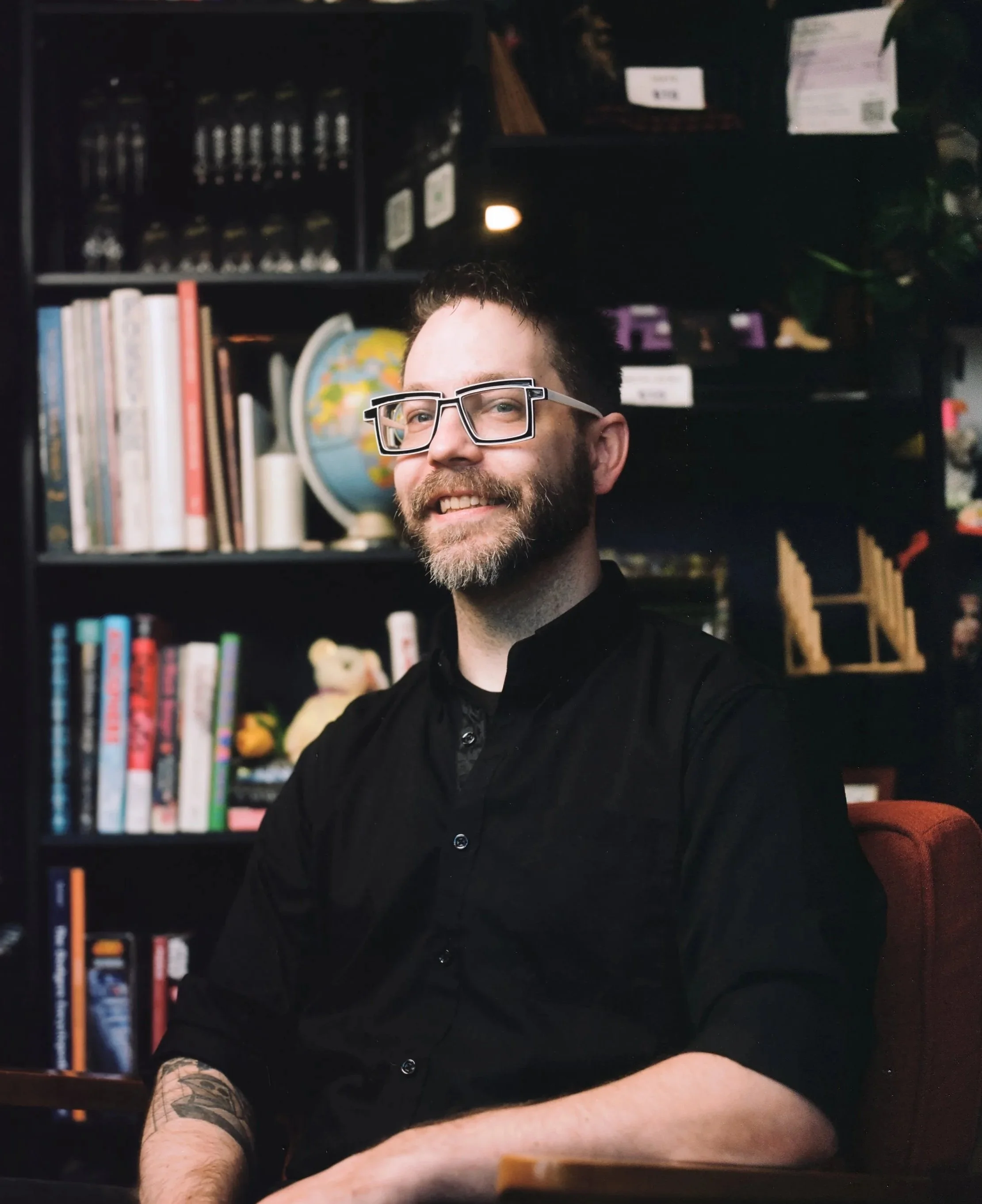 A smiling man with short dark hair, glasses, and a beard sitting in front of a bookshelf filled with books and a globe. He is wearing a black shirt and has a visible tattoo on his left arm. The background includes a dark bookshelf with items such as books, a globe, and decorative objects.