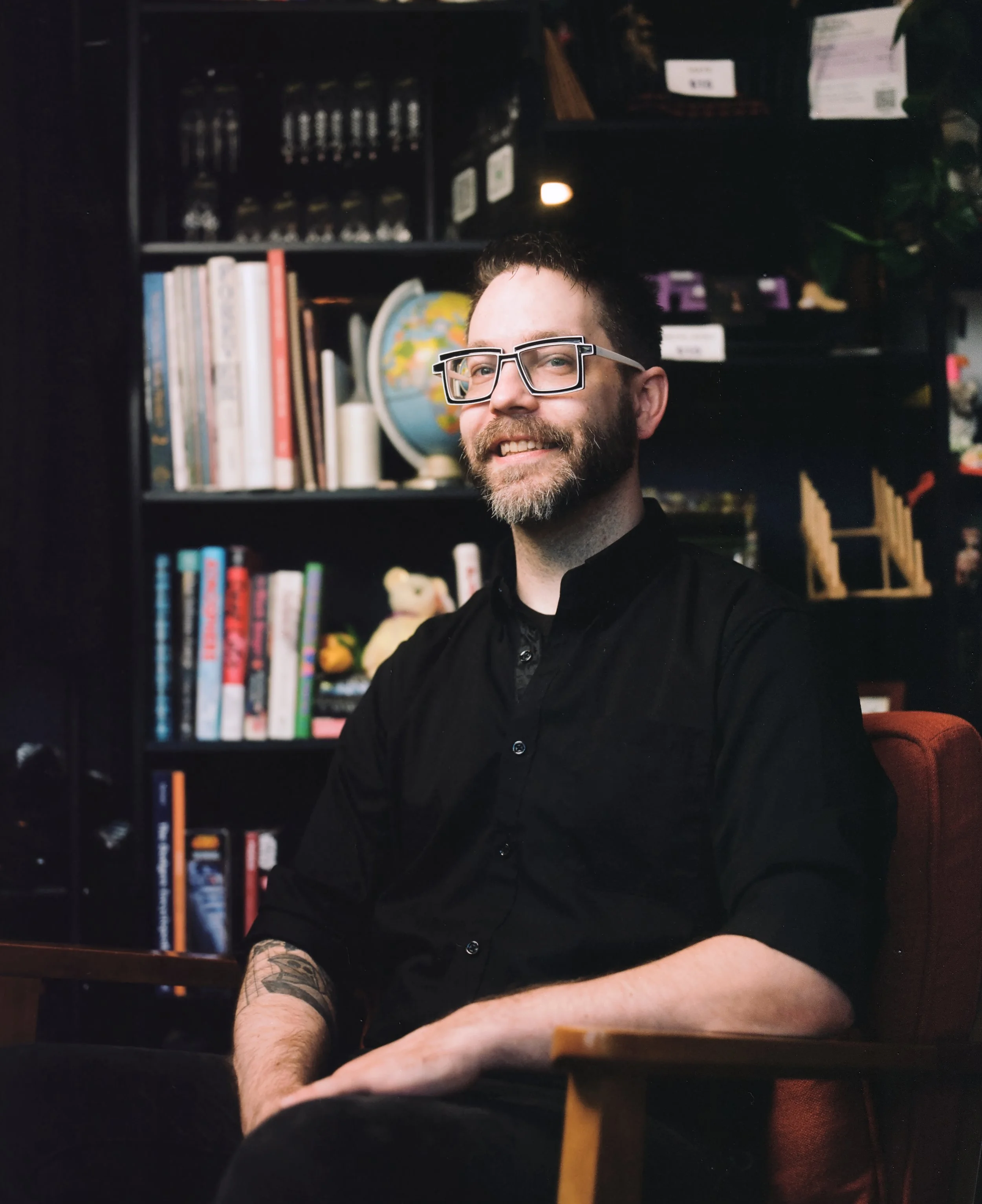 A man with glasses and a beard smiling, sitting at a table in a library or bookstore, with shelves of books in the background.